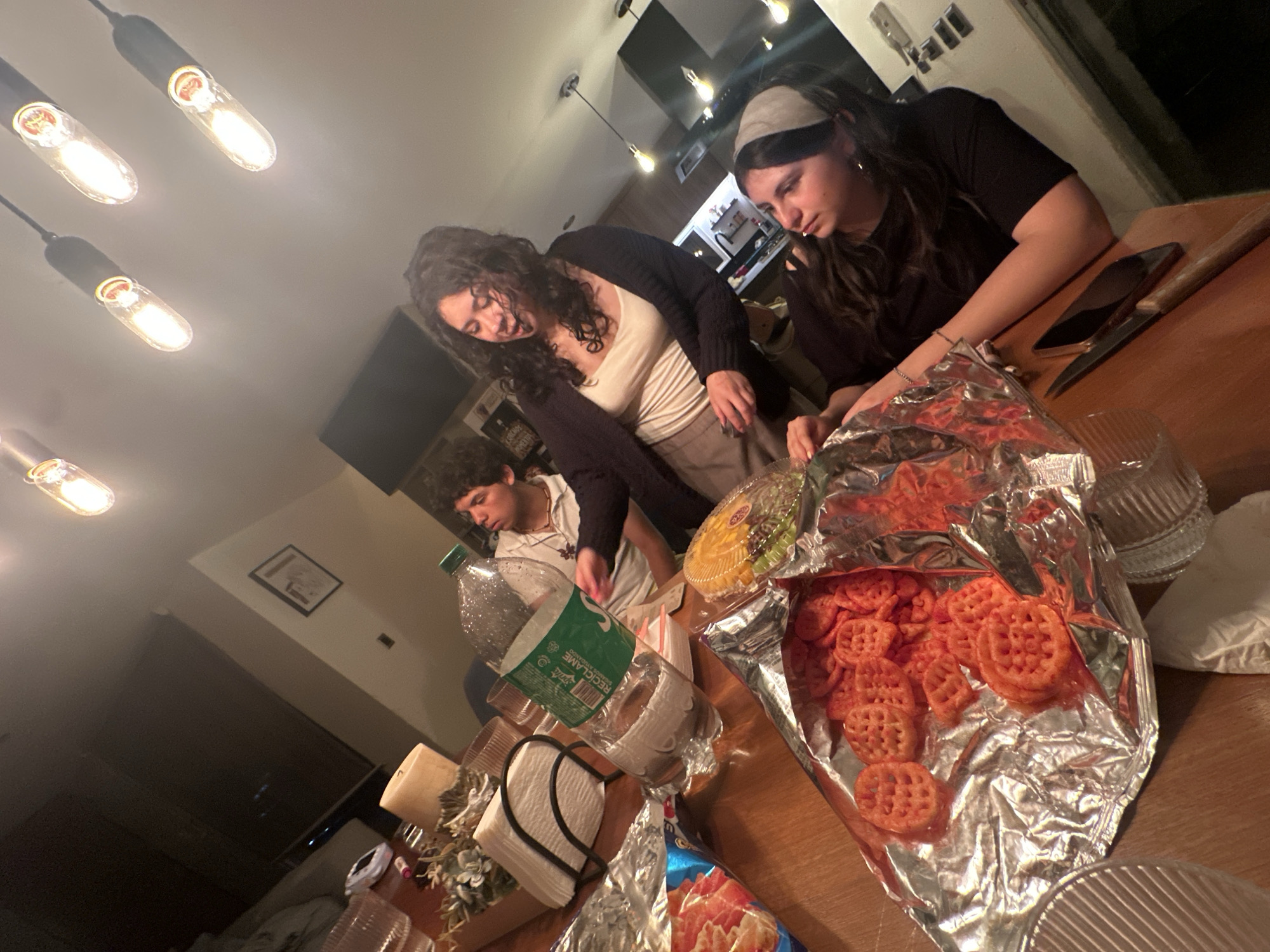 In San Bernardino Tlaxcalancingo, Mexico, a dimly lit interior scene captures three individuals gathered around a wooden table laden with snacks and drinks. A large bottle of "RECLAME" bottled water, green-labeled and partially filled, stands prominently. Two opened bags of orange, circular-shaped snacks are visible, one on aluminum foil and the other in a yellow plastic container. A napkin holder with white napkins and two lit candles are also on the table, suggesting a casual gathering or meal. The individuals, two women and one man, are dressed casually and appear to be interacting, with one woman gesturing while speaking. The lighting is provided by Edison-style pendant bulbs hanging from the ceiling, casting warm light on the scene. A kitchen counter and cabinets are visible in the background, along with a framed picture on the wall. The overall ambiance is informal and social.
