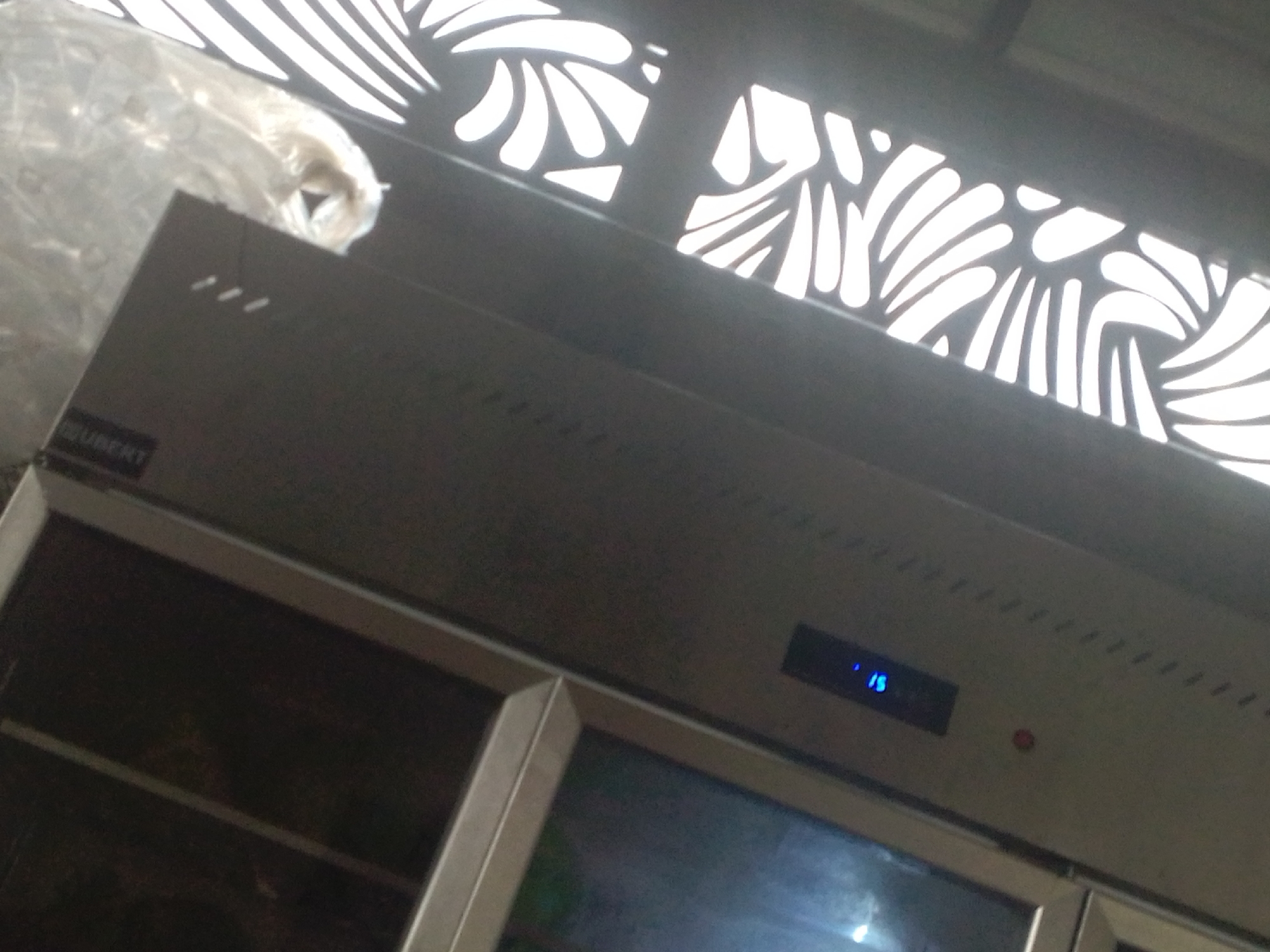 The image shows a stainless steel commercial refrigerator with a digital display showing "15". The refrigerator appears to be in a setting with a decorative metal grate overhead, allowing light to filter through. To the left of the refrigerator, a clear plastic bag, possibly containing an item, is draped over the top. The setting appears to be indoors, given the controlled lighting and the presence of appliances. There is no indication of people or a specific event taking place. The visible text on the refrigerator is "IZUBERT", suggesting the brand of the appliance. The location is identified as Howayi, Nigeria.