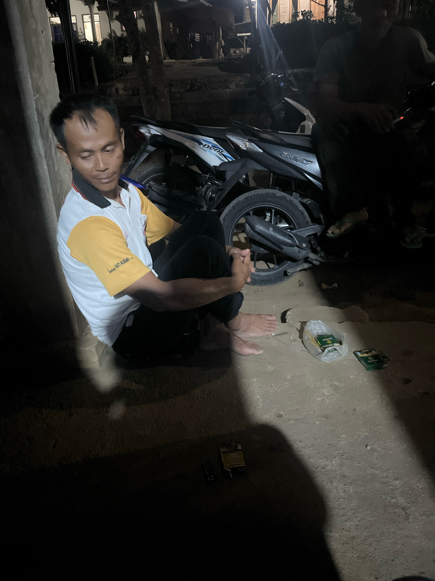 A man is sitting on the ground next to a motorcycle in what appears to be a residential area in Kebumen, Indonesia, at night. He is wearing a white polo shirt with yellow sleeves and black pants. He is looking down at his hands, which are clasped in front of him. There are two packs of cigarettes on the ground near him, one in a clear plastic bag and another loose. A second person is partially visible sitting on the motorcycle behind him, and a third person's legs are visible to the right of the motorcycle. The background shows a dimly lit building with windows and trees. The ground is a rough, concrete surface. The overall scene suggests a casual, informal gathering outdoors at night.
