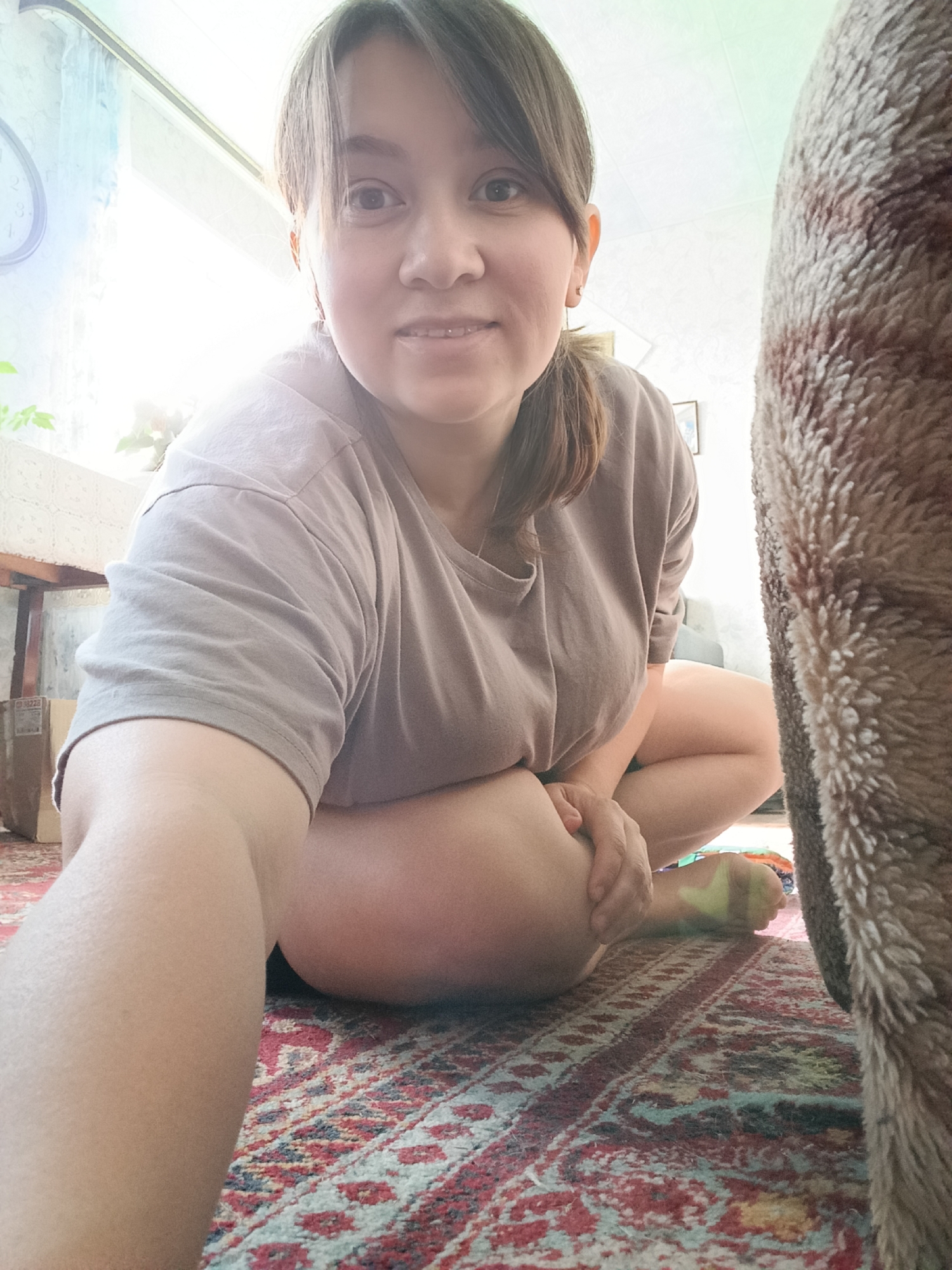 A young woman is captured from a low angle, facing the camera directly and smiling slightly. She has dark hair pulled back into a ponytail and is wearing a plain, light brown t-shirt. Her legs are bent and positioned in front of her, with her left arm resting on her knee. She is seated on a patterned rug with red, blue, and white motifs. To the right of the frame, a large, furry, brown and tan object, possibly a pet or a piece of furniture, occupies a significant portion of the right side. In the background, a wooden table and a window with sheer curtains are visible, with bright light streaming in from the left. A clock can be seen on the wall to the far left. The scene appears to be indoors, likely in a home environment, with no specific indicators pointing to the city of Каменное, Russia.