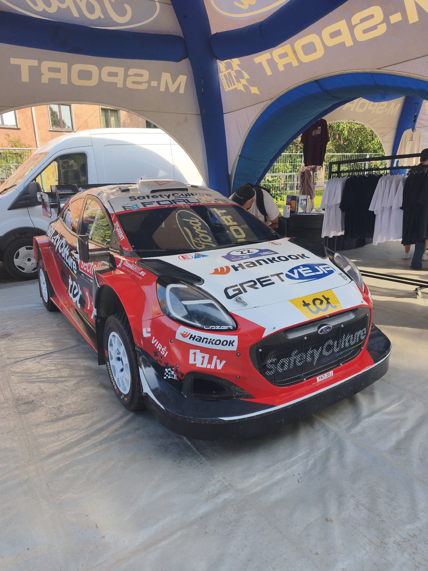 The image depicts a red and white rally car, a Ford Fiesta Rally3, parked on a concrete surface under a blue and white inflatable structure with "M-SPORT" branding. The car is adorned with various sponsor logos, including "SafetyCulture," "FORUM," "Hankook," and "GRETVEJA." A white van is visible behind the rally car. In the background, T-shirts are displayed on a rack, and people are present, one person appearing to work on the car and others standing around. The environment suggests an outdoor event or display, possibly at a motorsport venue. The presence of "GRETVEJA" and "VIRŠI" logos on the car indicates a connection to Latvia, and more specifically, could suggest a location within Riga, Latvia, where such motorsport events are held.