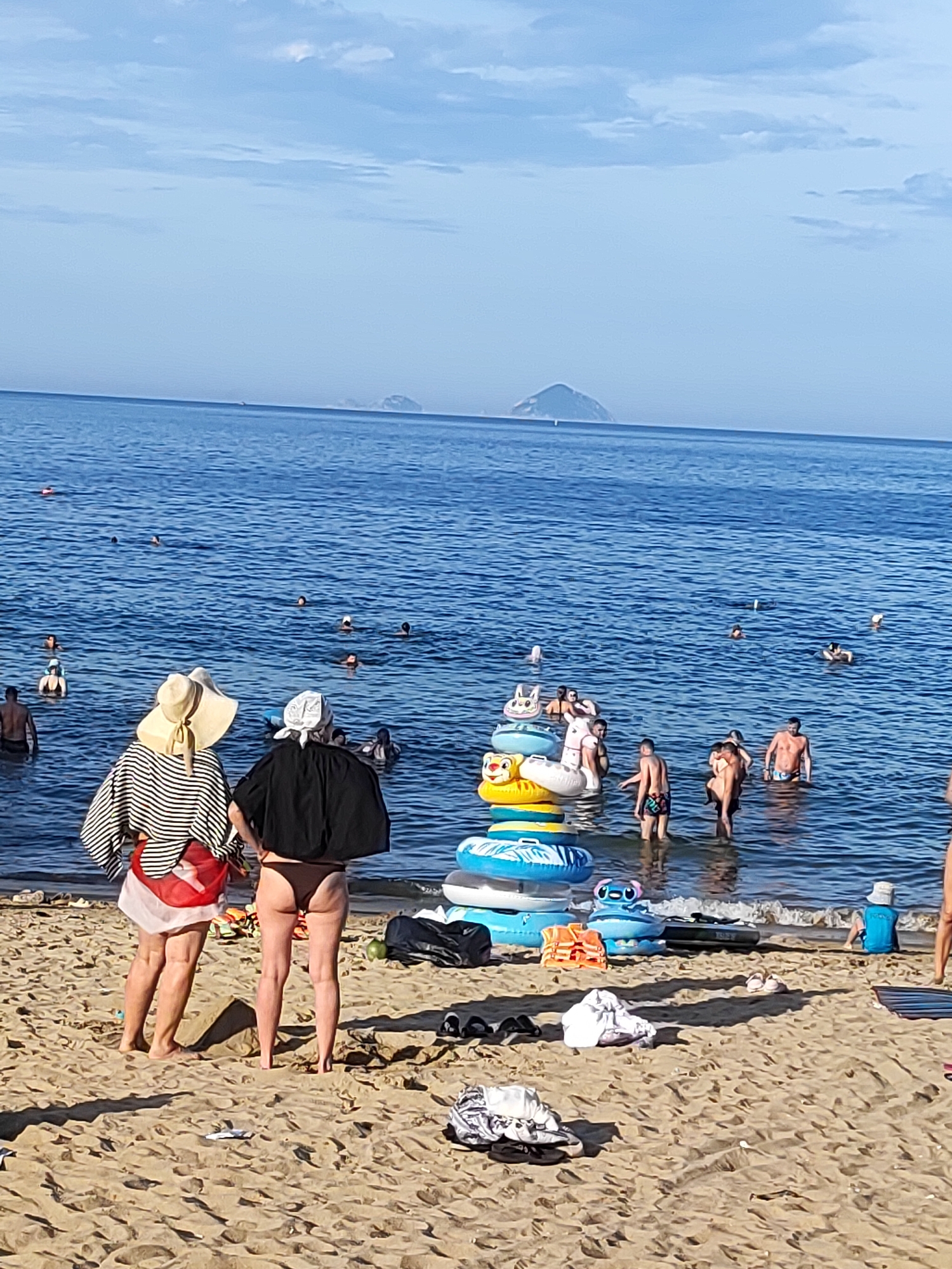 In Nha Trang, Vietnam, a beach scene unfolds with numerous individuals enjoying the water. The ocean is a vibrant blue, with a gentle wave line meeting the sandy shore. In the distance, two islands are visible on the horizon under a pale blue sky dotted with light clouds.
On the beach, two women stand facing the water. The woman on the left wears a wide-brimmed straw hat, a black and white striped long-sleeved shirt, and a red and white sarong. The woman to her right has a white headscarf and wears a black top and brown bikini bottoms. Their presence suggests they are either about to enter the water or have just exited.
The water is populated with many people, some swimming, others standing and conversing. A prominent feature in the middle ground is a stack of colorful inflatable rings, including a lion-faced ring and several blue and white ones. Among the inflatable rings, several people are standing in the shallow water.
Further out in the water, more individuals are visible, including a person in a blue life vest sitting on the sand near the water's edge on the right side of the frame. Scattered on the sand are various items such as bags, shoes, and discarded clothing, indicating ongoing beach activities. The overall atmosphere is one of leisure and recreation.