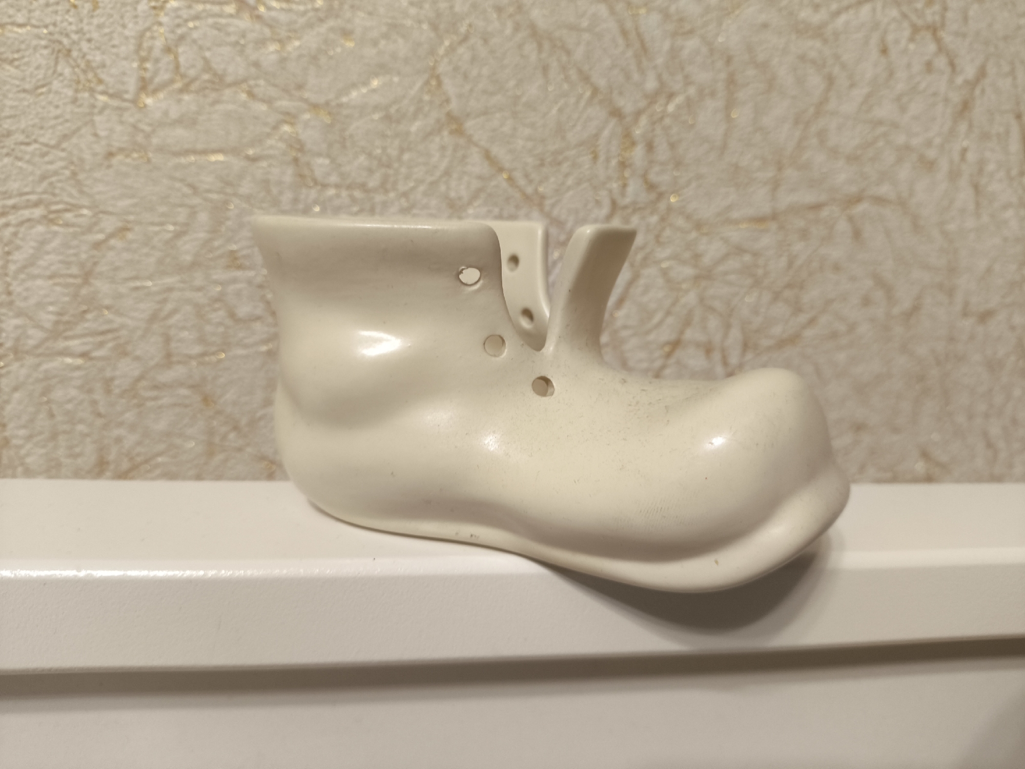 A small, off-white, ceramic shoe-shaped planter sits on a white, horizontally-oriented ledge. The shoe features rounded details and several small, circular holes. The background is a textured, light beige wallpaper with subtle gold patterns. The location is unknown and there are no indications that the image was taken in Moscow, Russia. No people or interactions are visible.