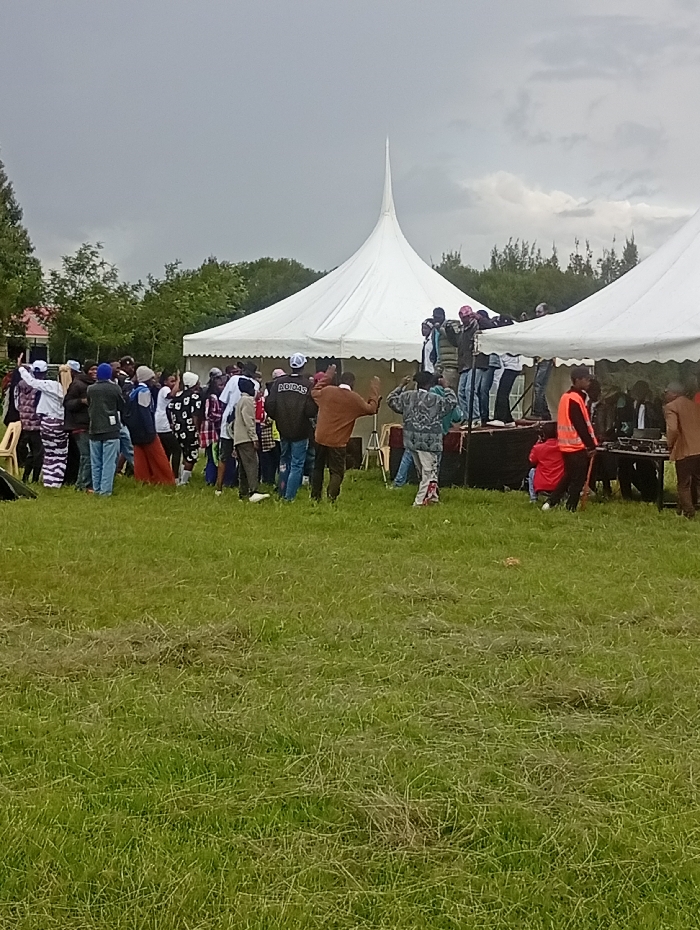The image depicts an outdoor gathering in Naro Moru, Kenya, under an overcast sky. A large group of people, predominantly adults and young adults, are congregated in a vast green field.
Two distinctive white event tents with tall, pointed peaks serve as central structures. Many individuals are clustered around and beneath these tents, especially near a raised, dark-colored stage or platform positioned in front of them. Some people are standing directly on the stage, while others are densely packed around its base, suggesting an active event such as a performance, speech, or communal activity. The crowd is casually dressed, with various jackets, shirts, and hats visible. One person's jacket clearly displays the "ADIDAS" logo on the back.
The background features a line of green trees and dense foliage, indicating a natural, possibly rural or semi-rural, outdoor setting. The sky is predominantly cloudy, suggesting either an overcast day or the possibility of rain. The foreground consists of natural green grass, showing some signs of foot traffic. The time of day is daytime, evidenced by the natural illumination. The overall scene conveys a sense of community engagement at an organized outdoor event.