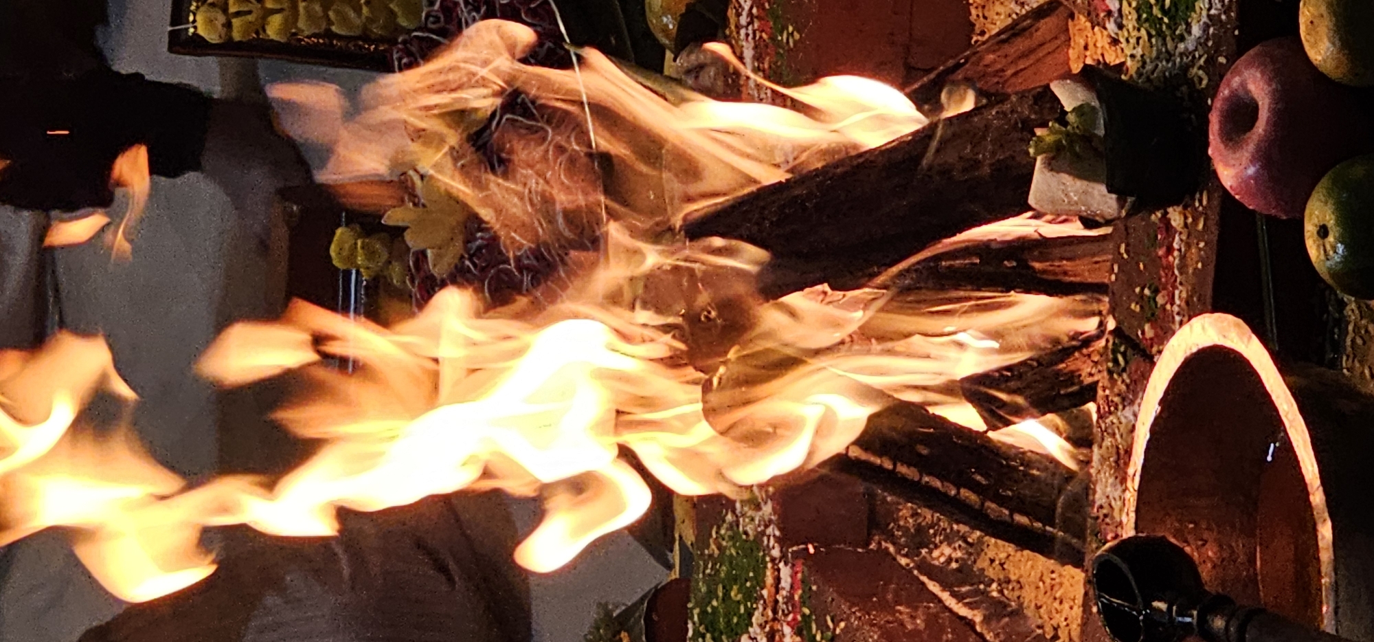 The image captures a vibrant ceremonial fire, likely part of a religious or cultural ritual in Denpasar, Indonesia. Bright orange and yellow flames dominate the central and left portions of the frame, indicating an active fire burning several pieces of dark wood or logs. The intense glow of the fire casts dynamic light on its surroundings, suggesting the event is taking place during the evening or night.
The fire is contained within a sturdy structure, possibly an altar or pit made of brick or stone, which is adorned with colorful, granular materials that appear to be powdered spices or ceremonial offerings. Various offerings are arranged around the fire: to the top-left, there are items that resemble yellow fruits or sweets, possibly on skewers, alongside hints of red floral arrangements. To the right, fresh fruits including a prominent red apple and a green, round fruit (possibly a mango) are visibly placed as offerings. In the bottom-right corner, a large, earthy-toned, open pot or bowl, along with a dark handle of a tool, is partially visible, likely used for the ritual.
A faint silhouette of a person can be discerned through the flames on the left side, indicating someone is actively tending to the fire or participating in the ceremony. The scene evokes a sense of solemnity and devotion, typical of a traditional spiritual event such as a Homa or Yajna, practices common in Balinese Hindu culture. There is no visible text in the image.