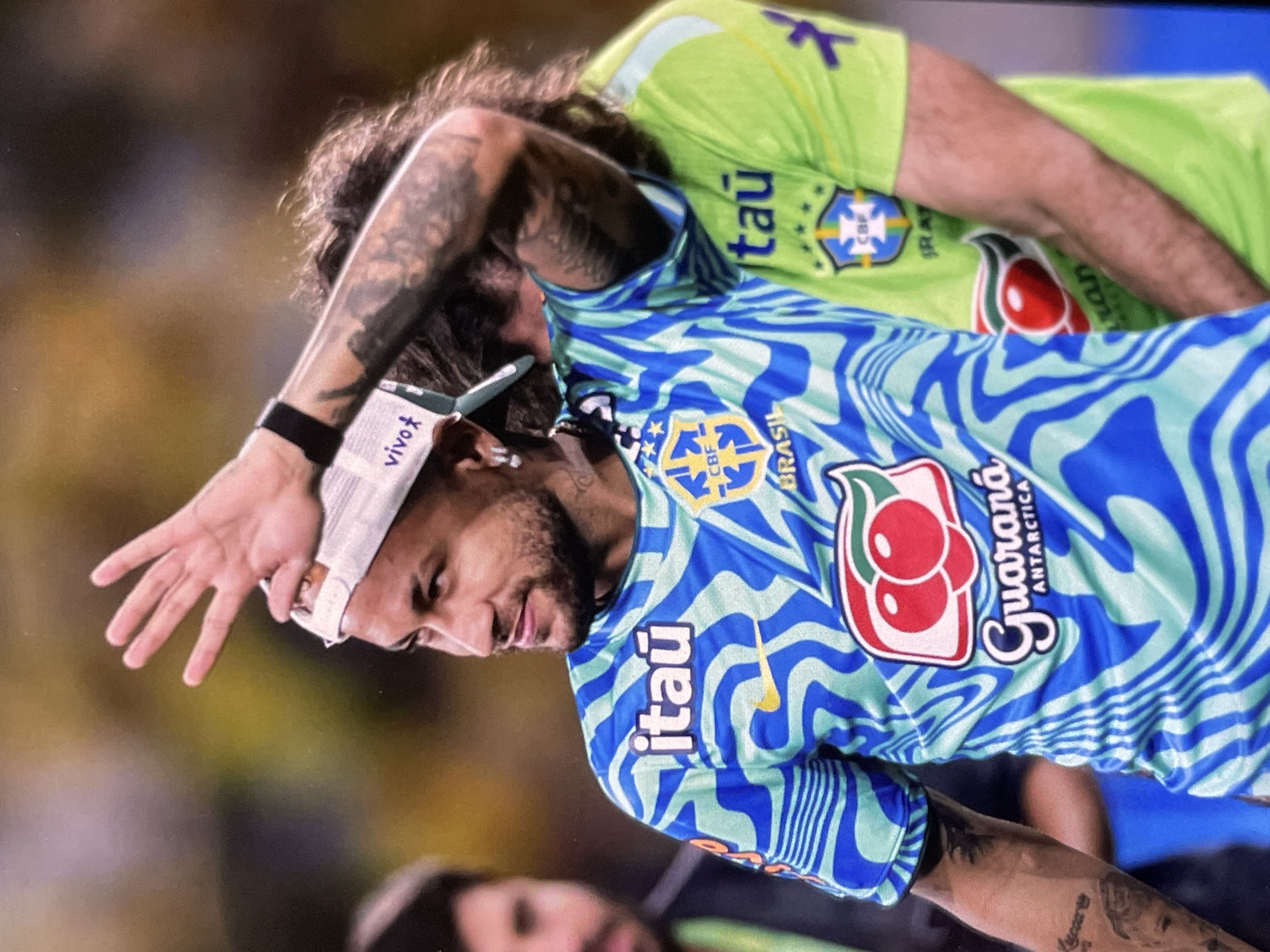 In the city of Unknown, Unknown, a male soccer player, identified as Neymar, is captured in a medium shot, his gaze directed to his right. He wears a distinctive teal and blue patterned jersey, emblazoned with the "Itaú" logo and the Brazilian national team crest featuring the CBF initials and stars. A red and white "Guaraná Antarctica" logo is prominently displayed on his chest. Neymar's left arm is raised, with his hand extended and fingers splayed, as if acknowledging an audience or signaling. He wears a white cap backward and a black wristband on his left arm. Tattoos are visible on his arms and neck. To his right, another player in a neon green jersey with a similar crest is partially visible, their arm resting on Neymar's shoulder. The background is blurred, suggesting a sporting event, with warm tones of yellow and brown indicating stadium lighting or an outdoor environment.