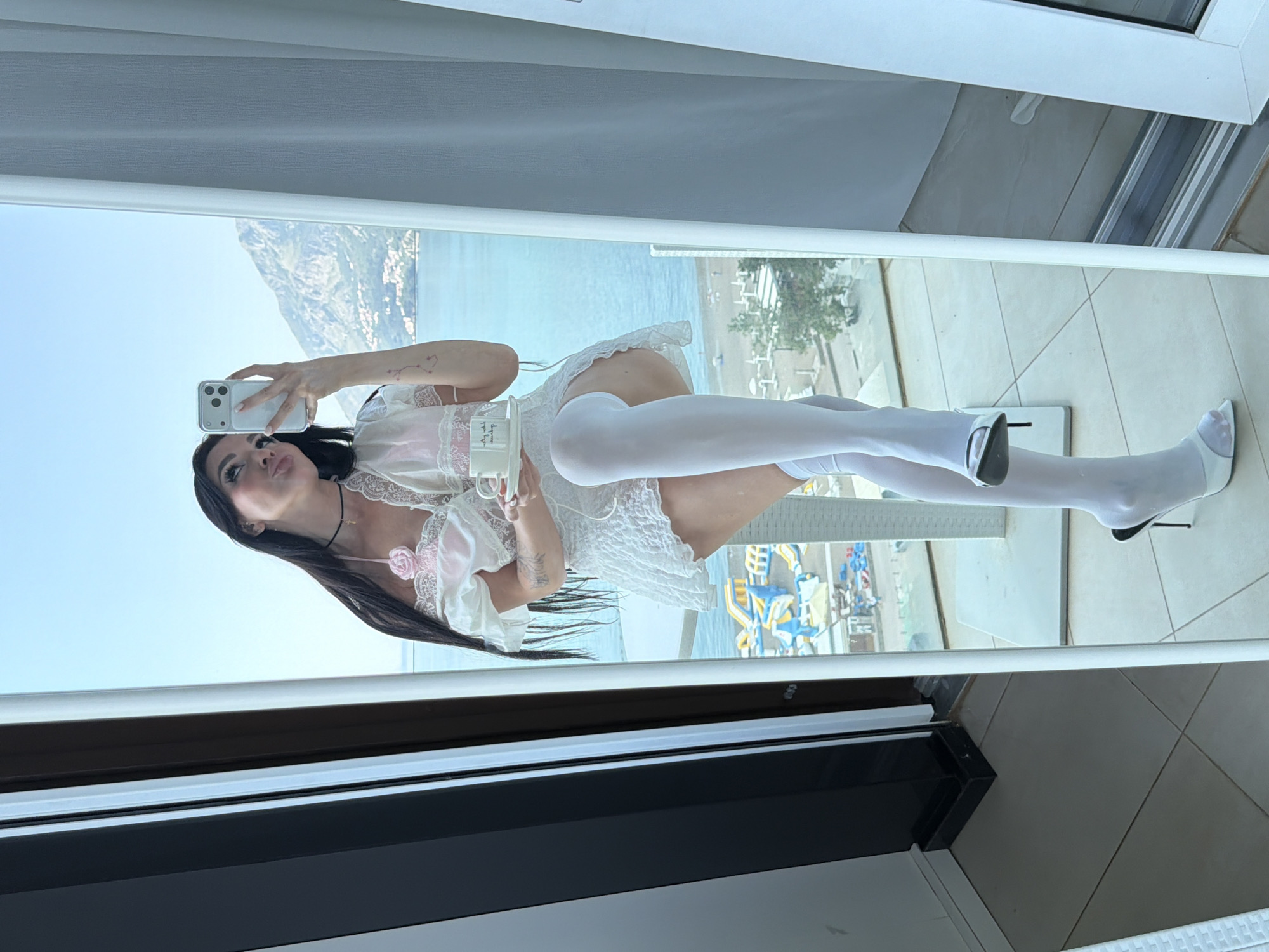 A woman is posing in front of a window in Boreti, Montenegro, using a mirror to take a selfie with her cell phone. She is wearing a white, lacy dress, white stockings, and white high heels. She is holding a mug in her left hand. Outside the window, there is a view of the sea, mountains, and a beach with sun umbrellas. The window frame is white.