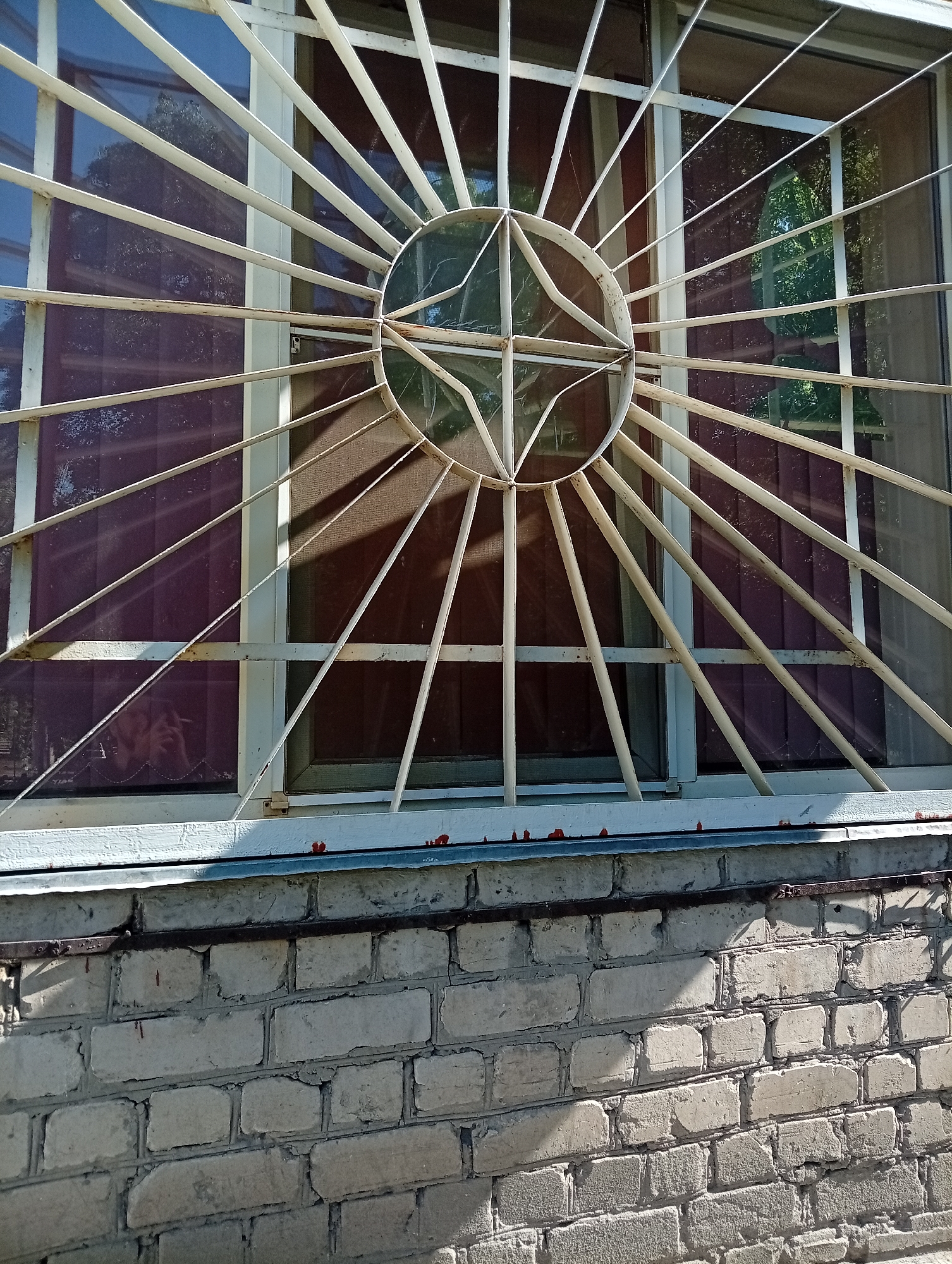 A close-up captures a window with a decorative, sunburst-patterned iron grille. The grille is painted white and features a circular center with radiating bars. The window behind the grille is divided into two panes, with what appears to be a screen visible in one pane. The window sits atop a concrete ledge, which in turn rests on a light-colored brick wall. The brick wall shows signs of age and weathering. Shadows are cast on the wall, suggesting sunlight. The photograph suggests an outdoor scene, likely a residential or commercial building.
