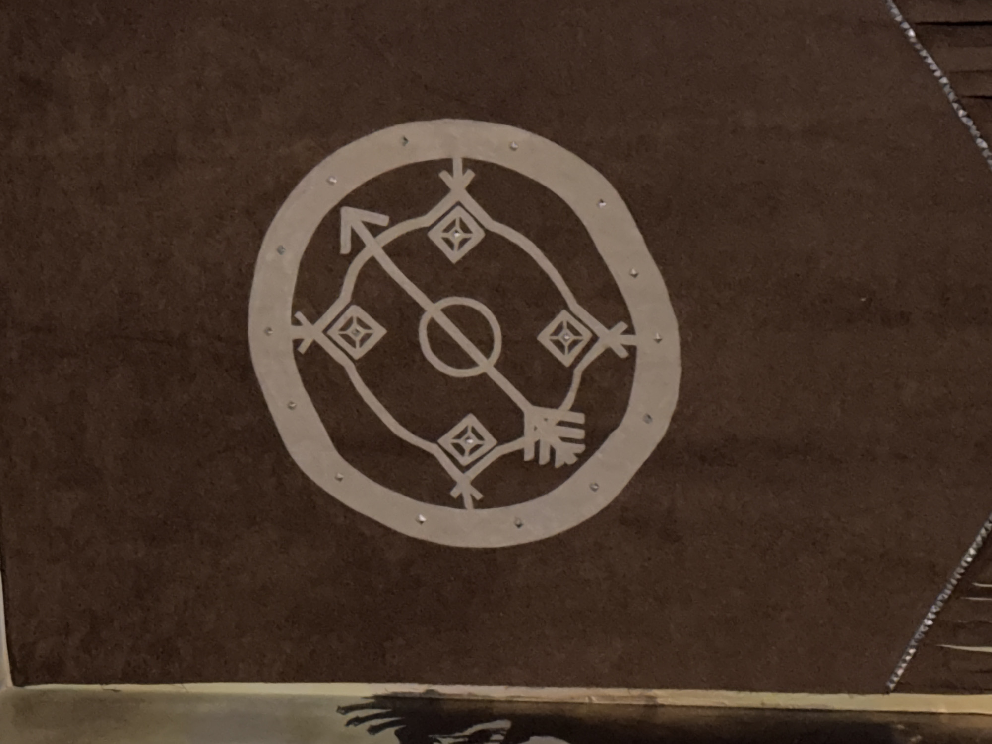 The image displays a close-up of a brown banner with a white symbol centered on it. The symbol is circular and contains an arrow piercing through a smaller inner circle. Four diamond shapes are positioned around the arrow, each containing a smaller diamond shape within. The outer edge of the circle is adorned with small, reflective dots. The banner has a decorative fringe at the bottom. The context provided indicates this banner is located in the city of Nyurba, Russia.