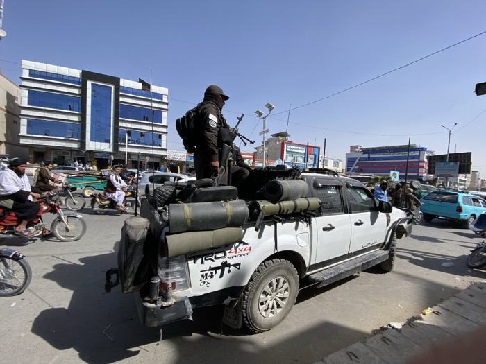 A white modified Toyota Hilux pickup truck, marked with "TRD 4X4 SPORT" and an M4 rifle graphic on its tailgate, occupies the foreground of a busy urban street in Kandahar City, Afghanistan. An armed male individual stands upright in the truck bed, wearing a dark uniform, tactical vest, backpack, and a dark face covering, holding an AK-style rifle. The truck bed is laden with several rolled green sleeping mats or tarps, camouflage bags, and other equipment. Another male in a blue shirt is visible in the passenger seat.
The scene depicts a major thoroughfare, likely a commercial district. To the left, a modern multi-story building with a white facade and prominent dark blue reflective glass windows stands prominently, consistent with structures found near central intersections like Shaheedan Chowk in Kandahar. Numerous motorcycles, ridden by males in traditional Afghan attire (shalwar kameez and turbans), navigate the street alongside the truck. Other vehicles, including sedans and a light blue hatchback, are visible in the surrounding traffic. Overhead power lines and streetlights equipped with solar panels are visible against a clear blue sky. The lighting indicates bright daylight conditions, casting distinct shadows on the asphalt road.