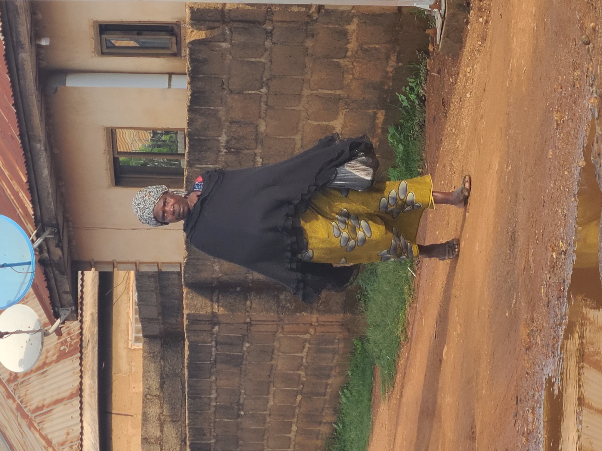 An elderly woman stands on a dirt path beside a brick wall in Jos, Nigeria. She wears a patterned head covering, a black shawl, and a yellow patterned dress. A building with visible windows and a satellite dish is behind her. The ground is wet with puddles, suggesting recent rain. The environment appears to be a residential area.
