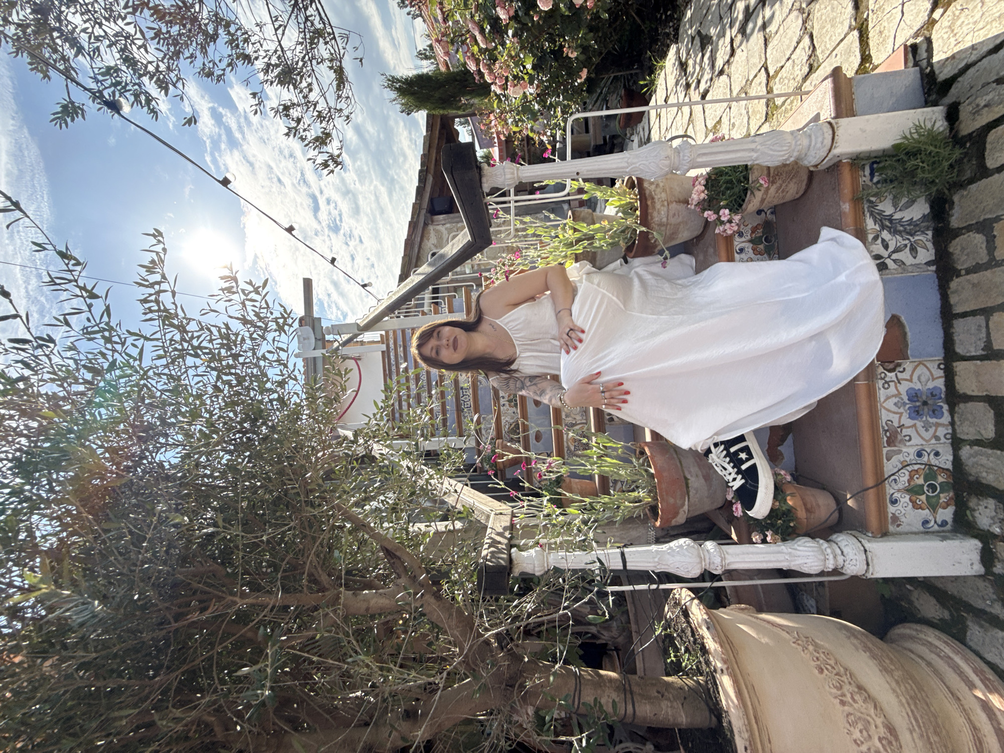 A woman is seated on a tiled outdoor staircase, wearing a long white dress and black sneakers. She has tattoos on her arms and is wearing rings. The staircase is adorned with potted plants, including an olive tree on the left. In the background, there is a building with a tiled roof and blooming pink roses. The sky is partly cloudy, and sunlight is visible. The scene appears to be in a garden or courtyard setting. There is no indication that this location is in Semogorskiy, Russia.