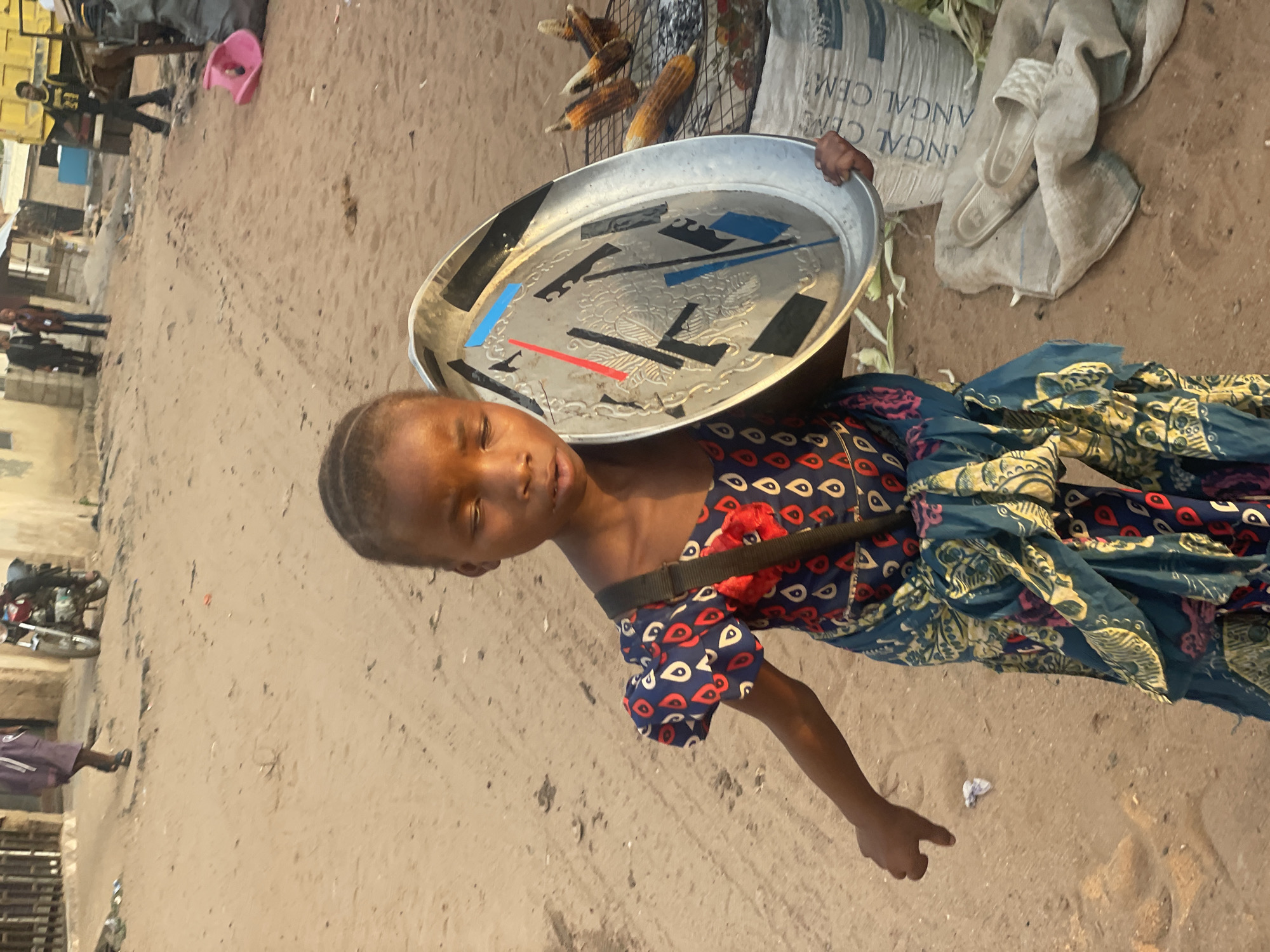 A young girl stands outdoors on a sandy surface, likely a street in Lokoja, Nigeria. She carries a large, round, aluminum tray strapped across her shoulder, with assorted objects, likely for sale, arranged on the tray. She wears a patterned blue and red dress and has a strap across her shoulder that holds the tray. Her right arm is extended, and she is pointing towards the ground.
Behind her are corn cobs arranged on a metal rack, a bag, and discarded fabrics. In the background, pedestrians, parked motorcycles, and buildings are visible. The lighting suggests it is daytime.