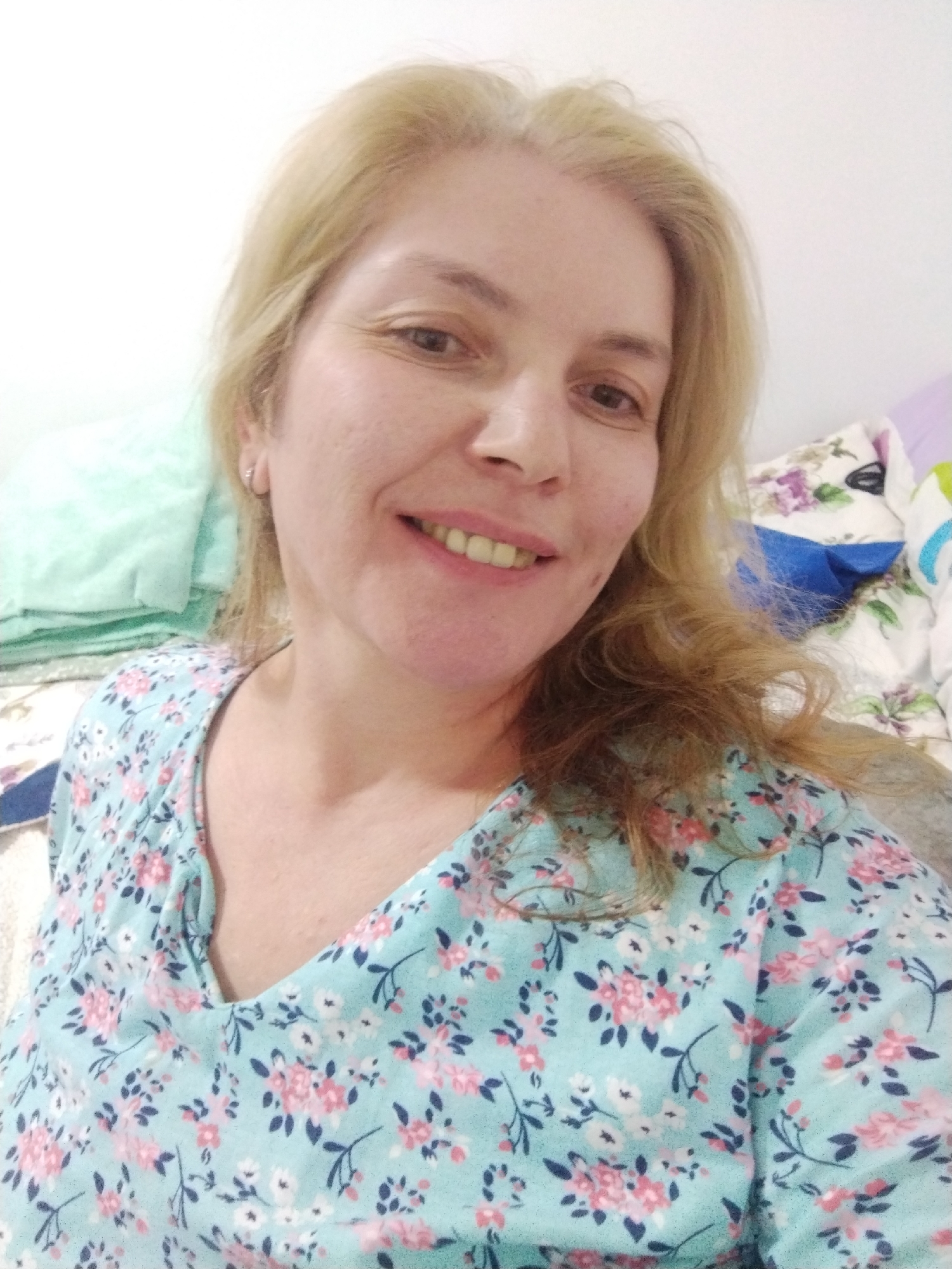 A medium close-up selfie of a light-skinned woman with blonde hair, fair skin, and a small nose ring smiles at the camera. She is wearing a light blue top with a floral pattern of pink and white flowers and green leaves. Her hair is medium length and slightly wavy, with some strands falling over her face. The background appears to be a bed with several pillows, one of which is light green and folded, and another with a floral pattern of purple and white flowers. The lighting is bright, suggesting an indoor setting. No specific location in Al Mas'oudi, United Arab Emirates can be determined from the image.