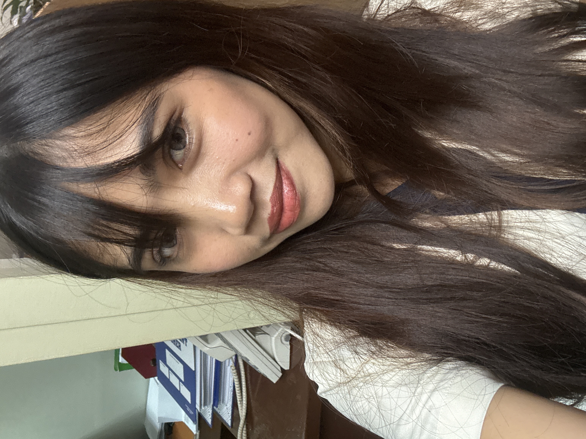 A close-up, indoor, waist-up photograph captures a woman with dark, straight hair framing her face. She is looking towards the camera with a slight smile and her lips are painted with a reddish-pink lipstick. Her eye makeup is visible, and her skin appears to have foundation and blush applied. She is wearing a light-colored, patterned shirt with a dark undershirt.
The background is out of focus but suggests an office environment. A desk is visible with stacks of papers and a telephone. To the left, a filing cabinet or shelf is partially seen, with some folders and documents. The lighting appears to be artificial.
There are no specific visual cues within the image that definitively place the location in Davao City, Philippines. The scene depicts a common indoor office setting that could be found in many urban environments.