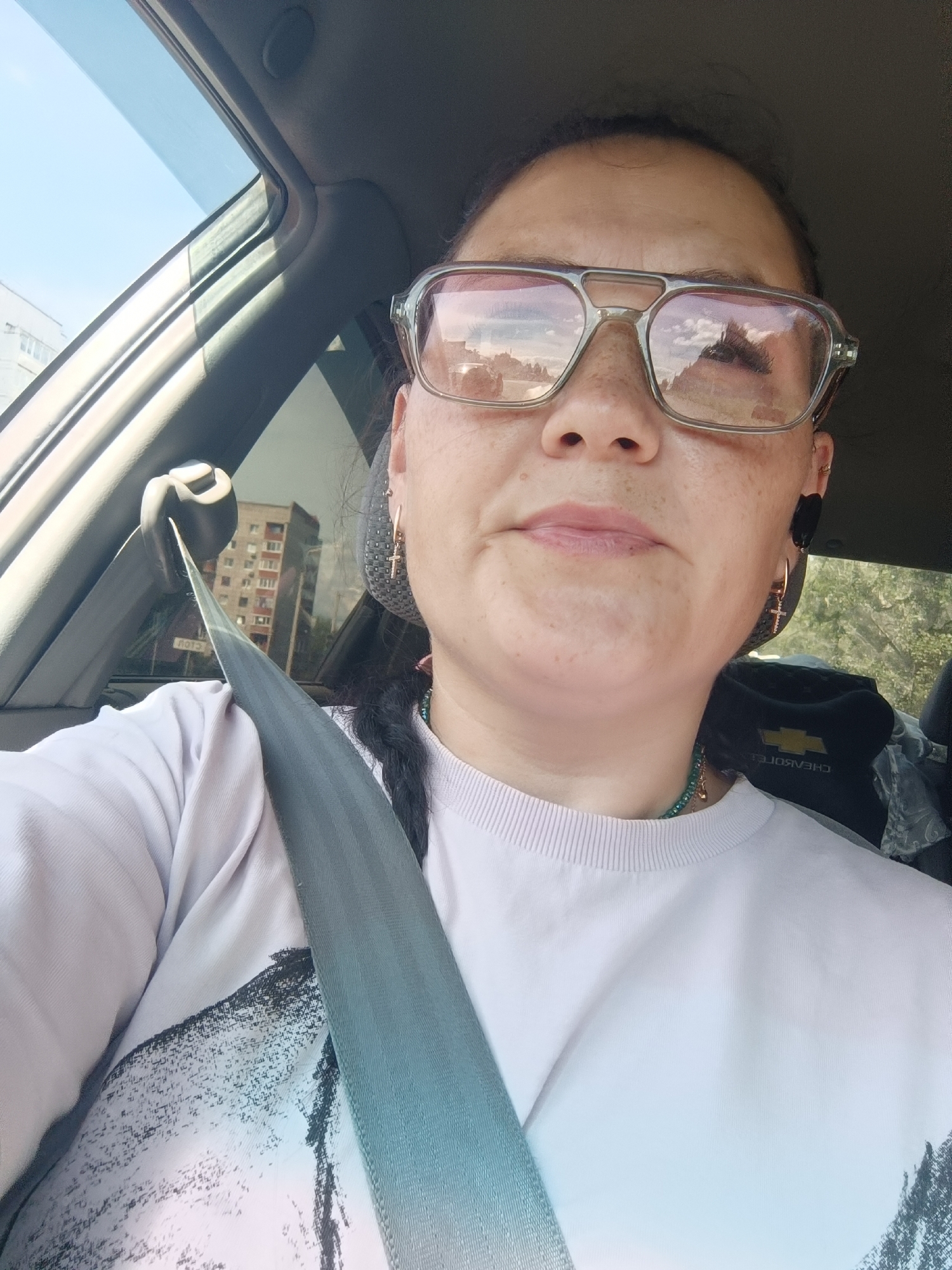 The image shows a close-up, waist-up, selfie-style photograph of a woman in a car, likely in Chaikovsky, Russia. The woman is wearing a light purple sweatshirt with a graphic print on the left side, a seatbelt across her chest, large-framed glasses with a pinkish tint, and a necklace. She has dark hair, styled in a braid, and is wearing earrings. The background shows a building visible through the car window, suggesting an urban environment. The sky is blue and clear. The interior of the car is visible, including part of the dashboard and the seatbelt mechanism. The overall lighting suggests it is daytime.