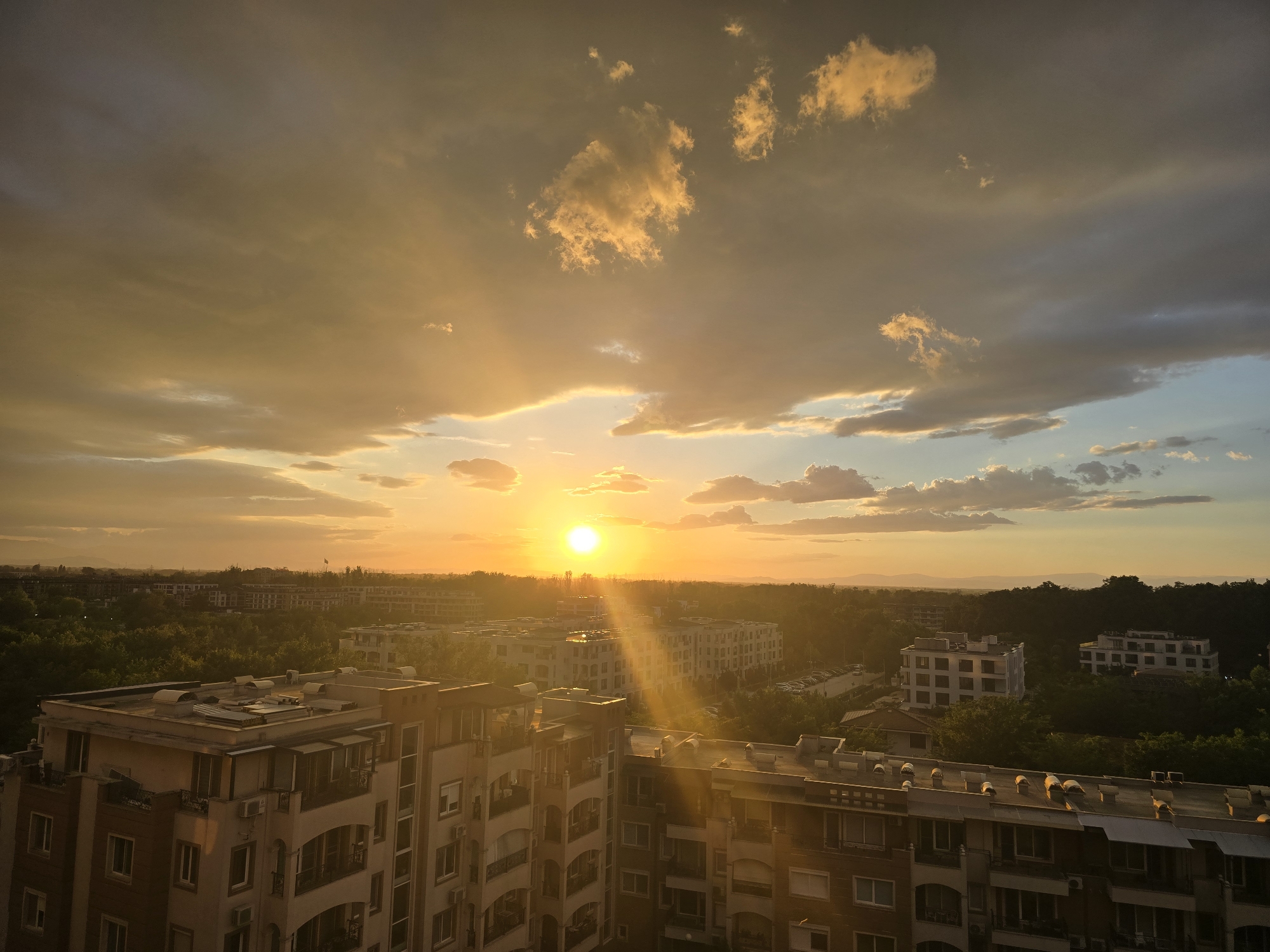 The image depicts a sunset over a city. The sun is positioned near the horizon, casting a golden glow across the sky and the buildings below. The sky is filled with clouds, some of which are illuminated by the setting sun. Below the horizon, a cityscape is visible, consisting of apartment buildings and trees. The location is the city of Unknown, Unknown.