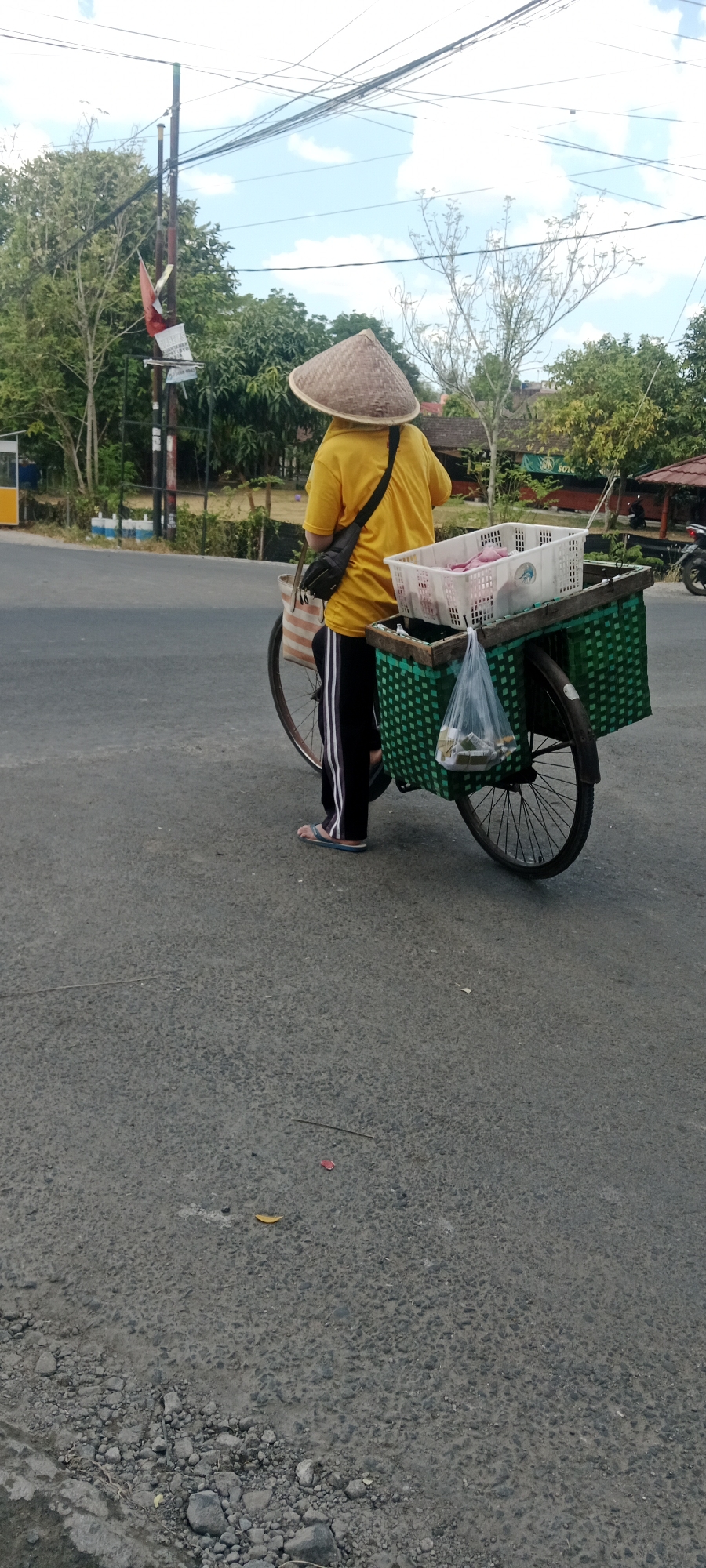 A person wearing a yellow shirt and a conical hat is standing next to a bicycle with baskets attached, on an asphalt road in Maguwoharjo, Indonesia. The bicycle is loaded with two white plastic baskets, one on top of a green woven basket. The top basket contains what appears to be raw meat. The green woven basket has a clear plastic bag hanging from it, filled with small packaged items, possibly snacks or cigarettes. The person is wearing black pants with white stripes and flip-flops. Behind the person, trees and power lines are visible against a partly cloudy sky. A pole with a red and white flag is also present. The overall scene depicts a street vendor preparing to sell goods.