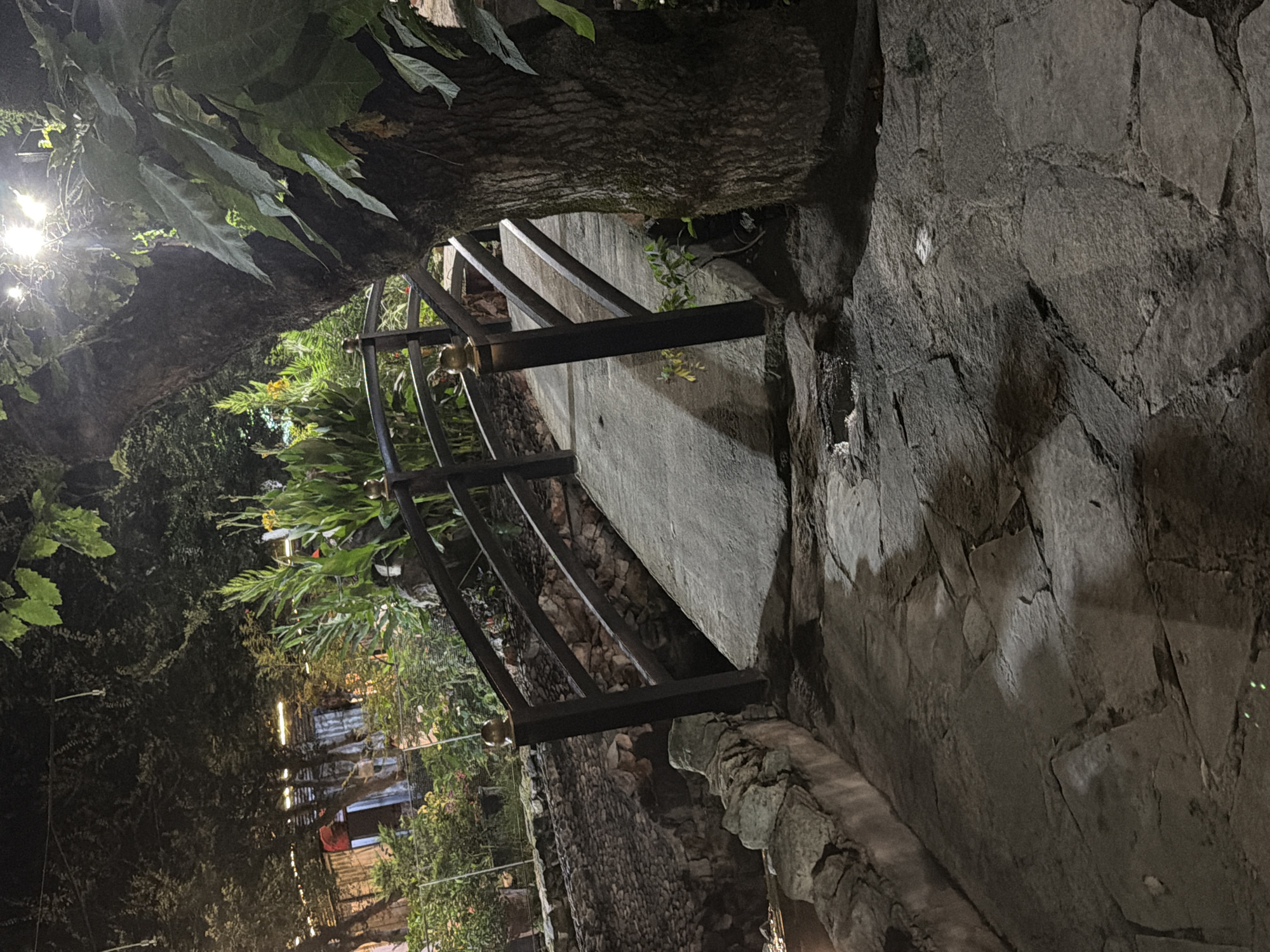The image depicts a nighttime scene featuring a small pedestrian bridge constructed of concrete and wood. The bridge has dark wooden railings with brass accents. It spans a narrow, stone-lined channel. The path leading to the bridge is paved with irregular flagstones, also illuminated by artificial light. Adjacent to the bridge and path is a large tree with a textured trunk, its leaves partially obscuring a bright overhead light source. Beyond the bridge, lush green foliage and buildings with illuminated windows are visible. The scene appears to be in an outdoor, possibly garden or park-like setting.