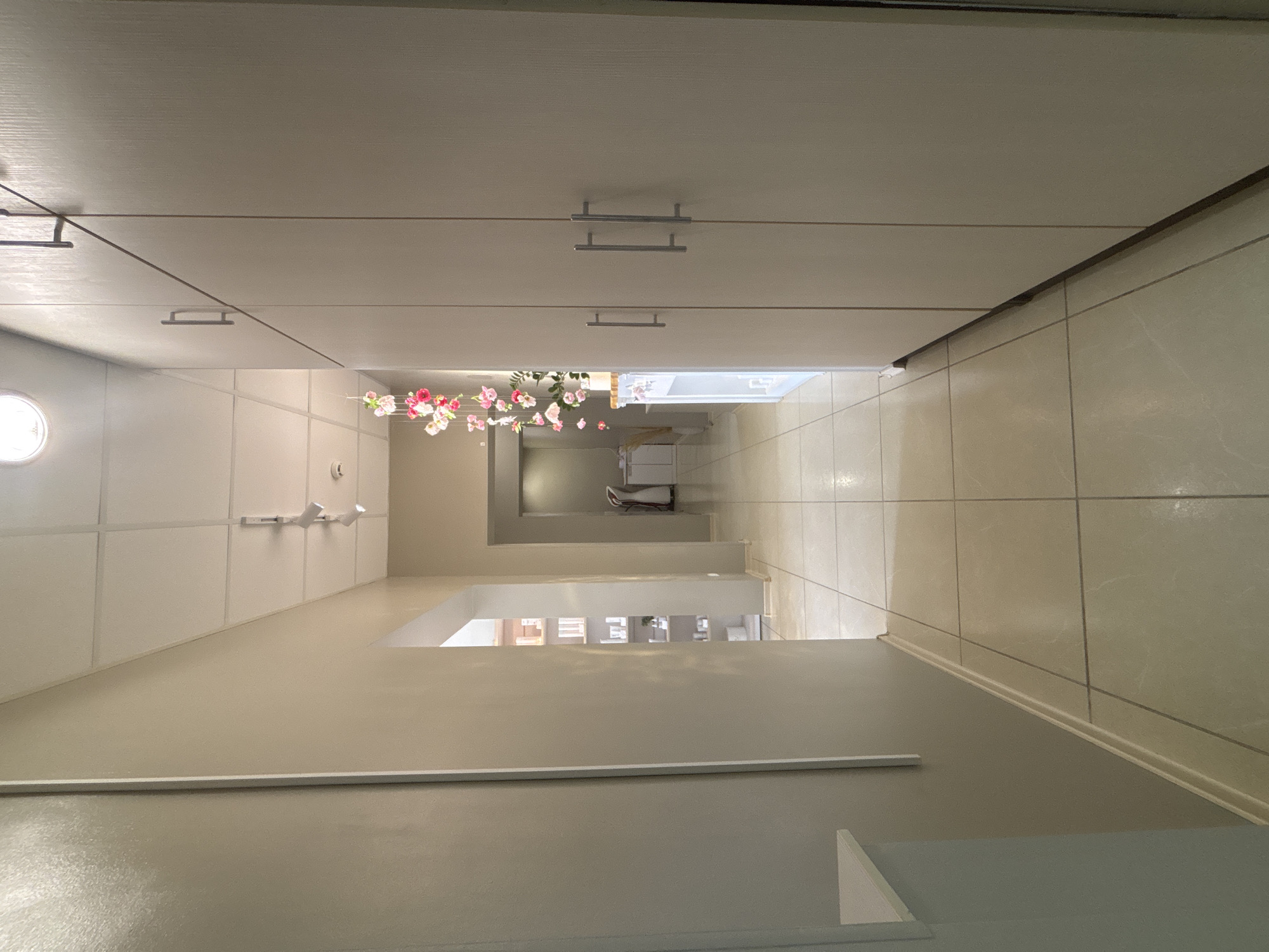 A long, narrow hallway is seen from a low angle. Above the hallway are cabinets with silver handles. The walls are light beige. At the end of the hallway is a bathroom with white cabinets. The floor is tiled. A string of artificial pink flowers hangs from the ceiling of the hallway. The image does not contain information suggesting it is located in Vsevolozhsk, Russia.