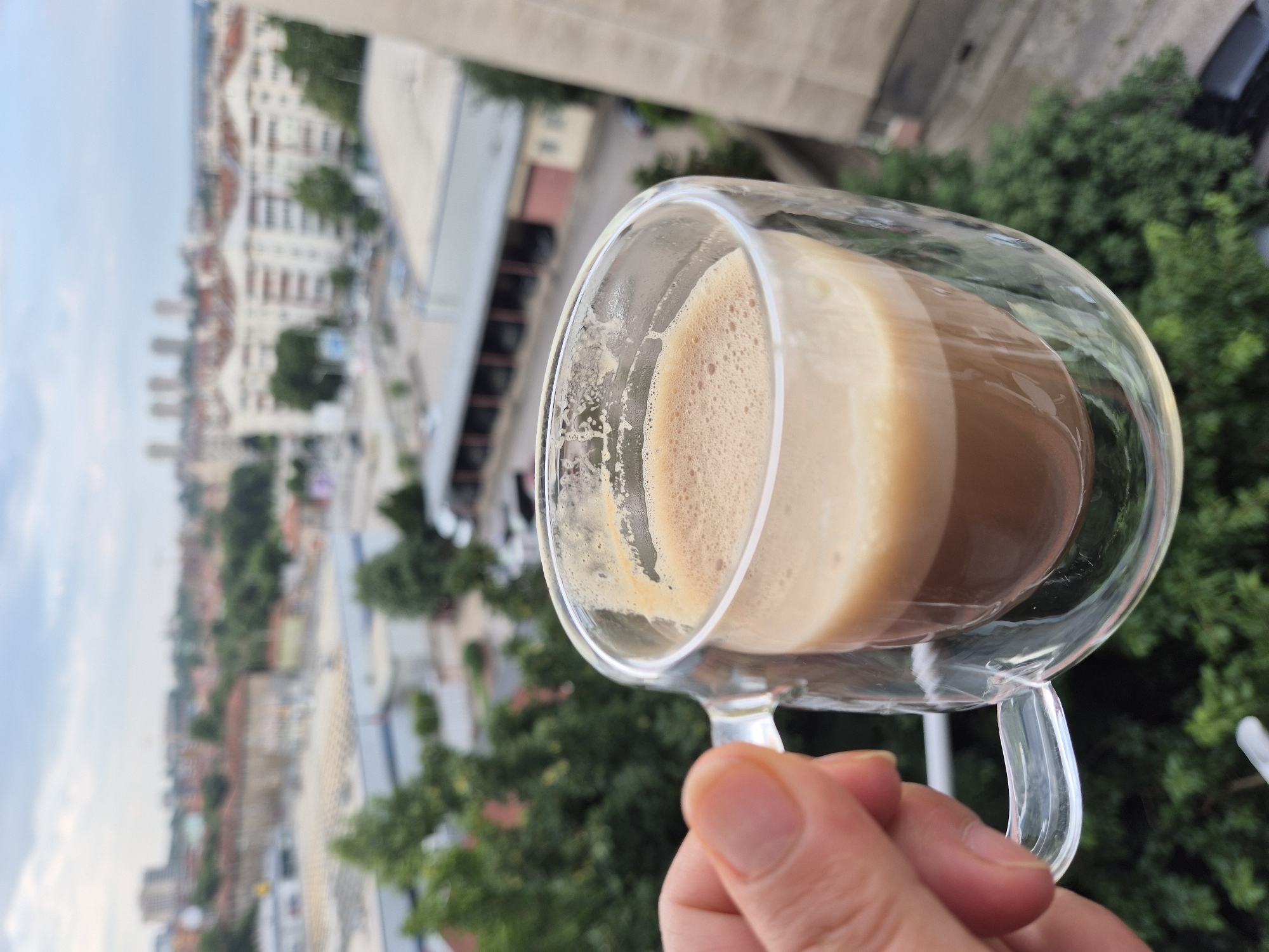 A hand is holding a double-walled glass mug filled with coffee, with a foamy top layer. The mug is held in the foreground, slightly to the left of the frame. In the background, a blurred cityscape is visible under a cloudy sky. The cityscape includes buildings with red roofs, residential buildings, and some commercial structures. Green trees are also interspersed within the urban landscape. The overall scene suggests a person enjoying a coffee break with a view of the city.
