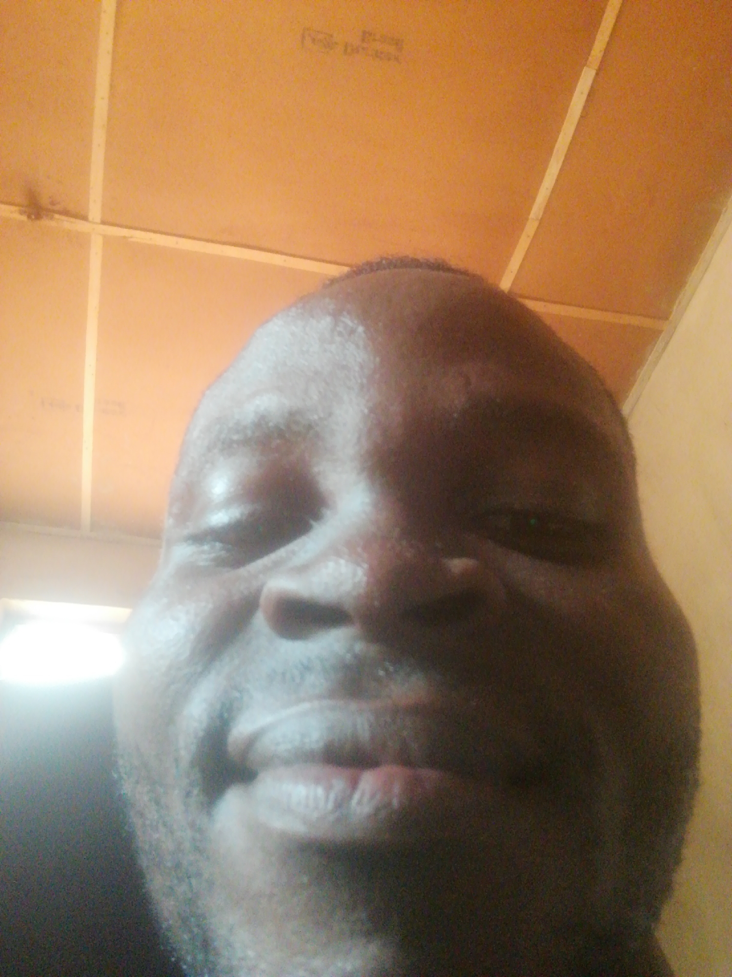 A close-up selfie of a Black man with a bald head, looking down at the camera. The background is a brown ceiling with white support beams. The man's face is sweaty and lit from the left. Location: Unknown, Unknown.