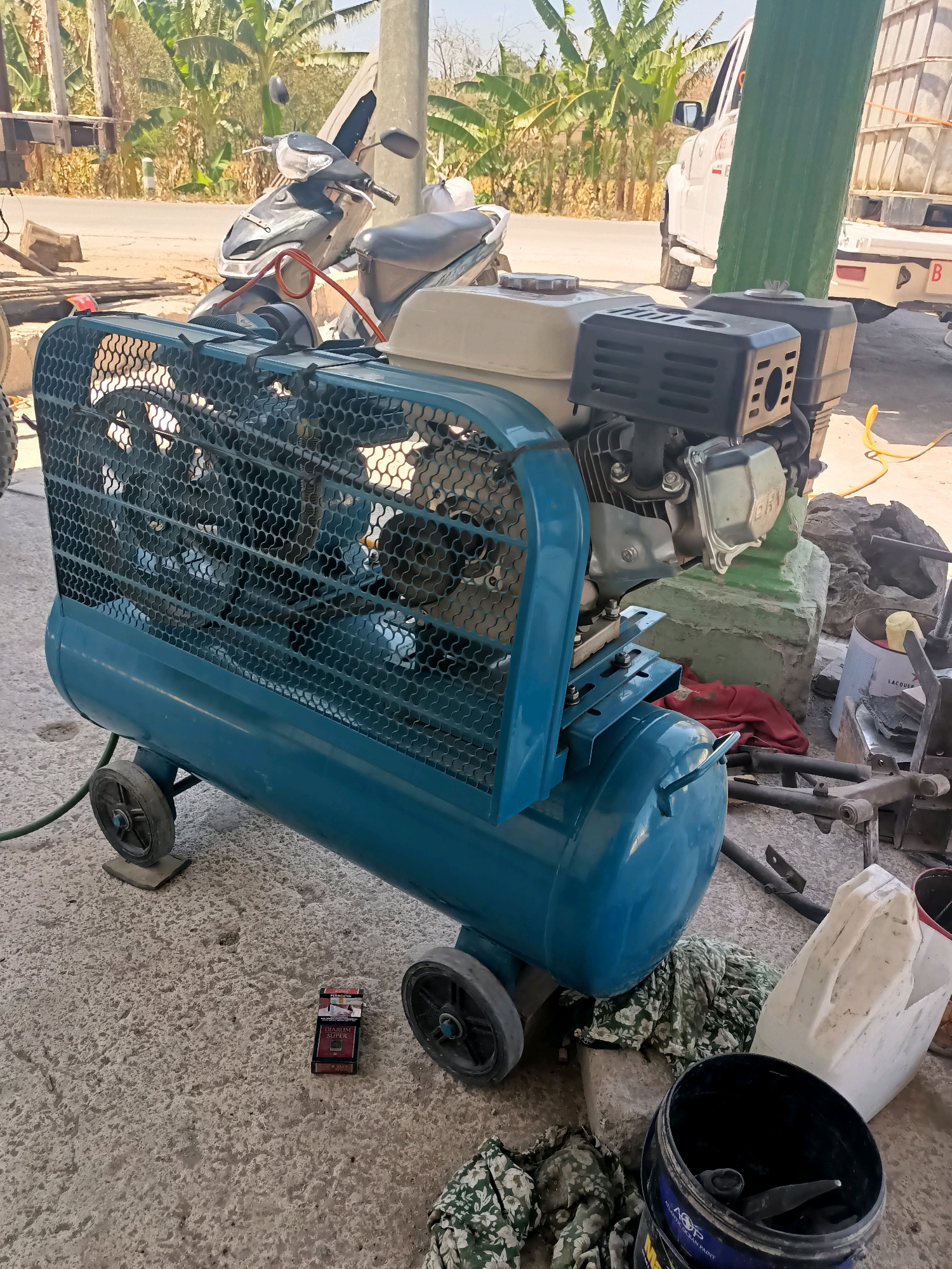The image depicts an air compressor with a petrol engine in an outdoor setting in Sumber Bendo, Indonesia. The air compressor is blue and sits on a concrete surface, supported by two wheels in the front and two in the rear. It has a metal mesh guard covering the engine and fan. In the background, there are several motorcycles parked, a white truck, and vegetation including banana trees. Various objects are scattered around the scene, such as rocks, fabric, and containers, suggesting a workshop or storage area. A pack of cigarettes is placed on the concrete in front of the air compressor. The lighting suggests it is daytime.