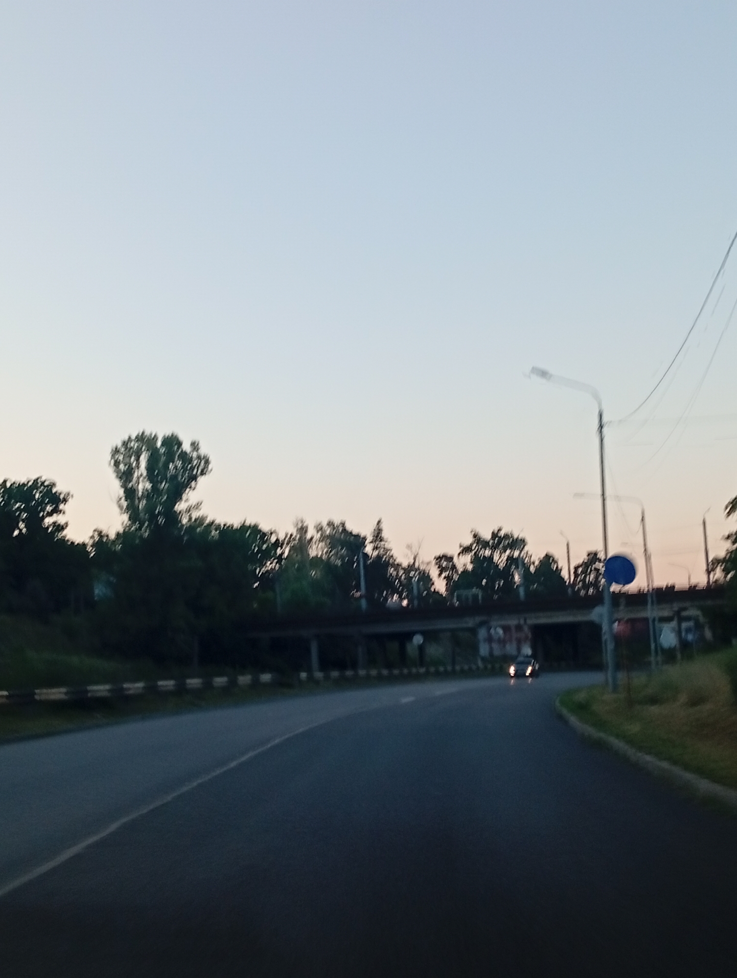 The image depicts an outdoor scene, likely during twilight or dawn, with a clear, pale sky dominating the upper portion. A road curves through the frame, with a light-colored line marking its edge on the left. The road surface is dark asphalt. On the left side of the road, a raised embankment is visible, topped with a dark-colored barrier that has a series of reflective white horizontal stripes. Trees, some appearing dense and dark, line the embankment. A tall, somewhat bare tree with a distinctive canopy stands out against the sky.
In the mid-ground, an overpass or bridge structure crosses the scene, supported by columns. Beneath the bridge, some graffiti or markings are partially visible on the concrete. A few streetlights are present along the right side of the road. One prominent streetlight is tall and slender, with a curved arm holding a light fixture. A blue circular sign is attached to a pole near this streetlight. Power lines are strung across the sky, originating from the upper right.
Further down the road, a single car is visible, its headlights illuminated, appearing to be traveling away from the camera. The right side of the road features a grassy verge and more vegetation. The overall impression is of a road in a semi-urban or suburban area at the edge of daylight.
