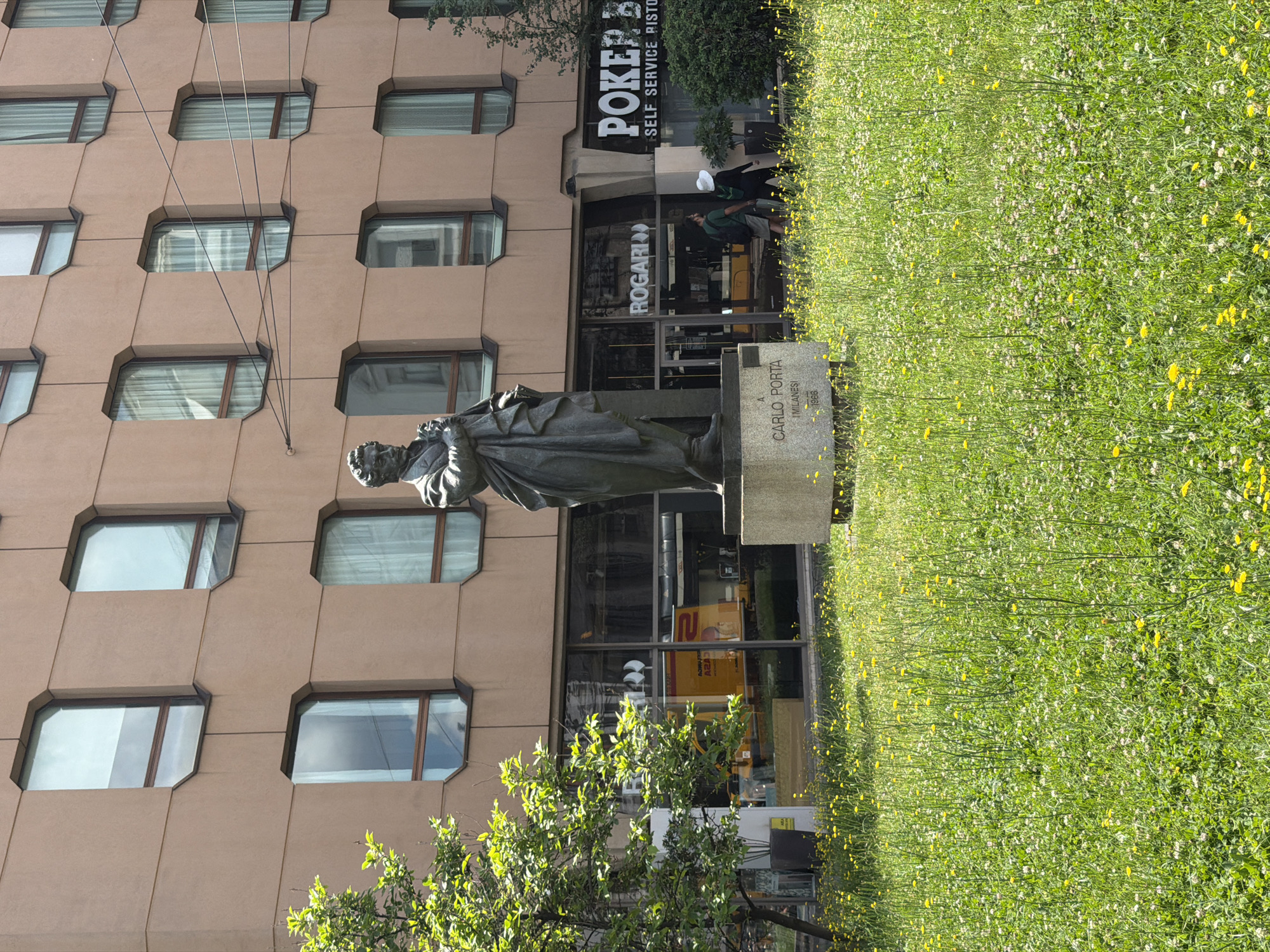 The image displays an urban scene featuring a statue in front of a modern building, bordered by a vibrant green lawn. The picture is taken during the daytime under sunny conditions, indicated by the bright lighting and clear shadows.
In the foreground, a verdant lawn densely covered with small yellow and white wildflowers, resembling dandelions and clover, stretches across the lower part of the frame. To the left, a bronze statue of a man, possibly a poet or intellectual, is depicted in a long, draped garment, standing on a light-colored stone pedestal. The pedestal bears the inscription: "A CARLO PORTA MILANESE 1986," identifying the subject as Carlo Porta, a Milanese poet.
Behind the statue and lawn stands a multi-story building with a light brown or tan facade. The building features an array of distinctive octagonal windows with dark frames, arranged in a grid pattern. At ground level, the building has a glass storefront, reflecting parts of the surroundings. Several signs are visible on the storefront, displaying text such as "PÖKER B..." (likely "PÖKER BAR"), "- SELF SERVICE RISTO..." (likely "SELF SERVICE RISTORANTE"), and "ROGARI>>". A few pedestrians are visible near the storefront, including one person wearing a white hat and another in a green shirt.
A small tree with bright green leaves is situated in front of the building, partially obscuring the lower part of the statue and the storefront. Overhead, several electrical or utility wires traverse the scene, adding to the urban feel. The overall setting suggests a public square or park area within Milan, Italy, honoring a local historical figure.