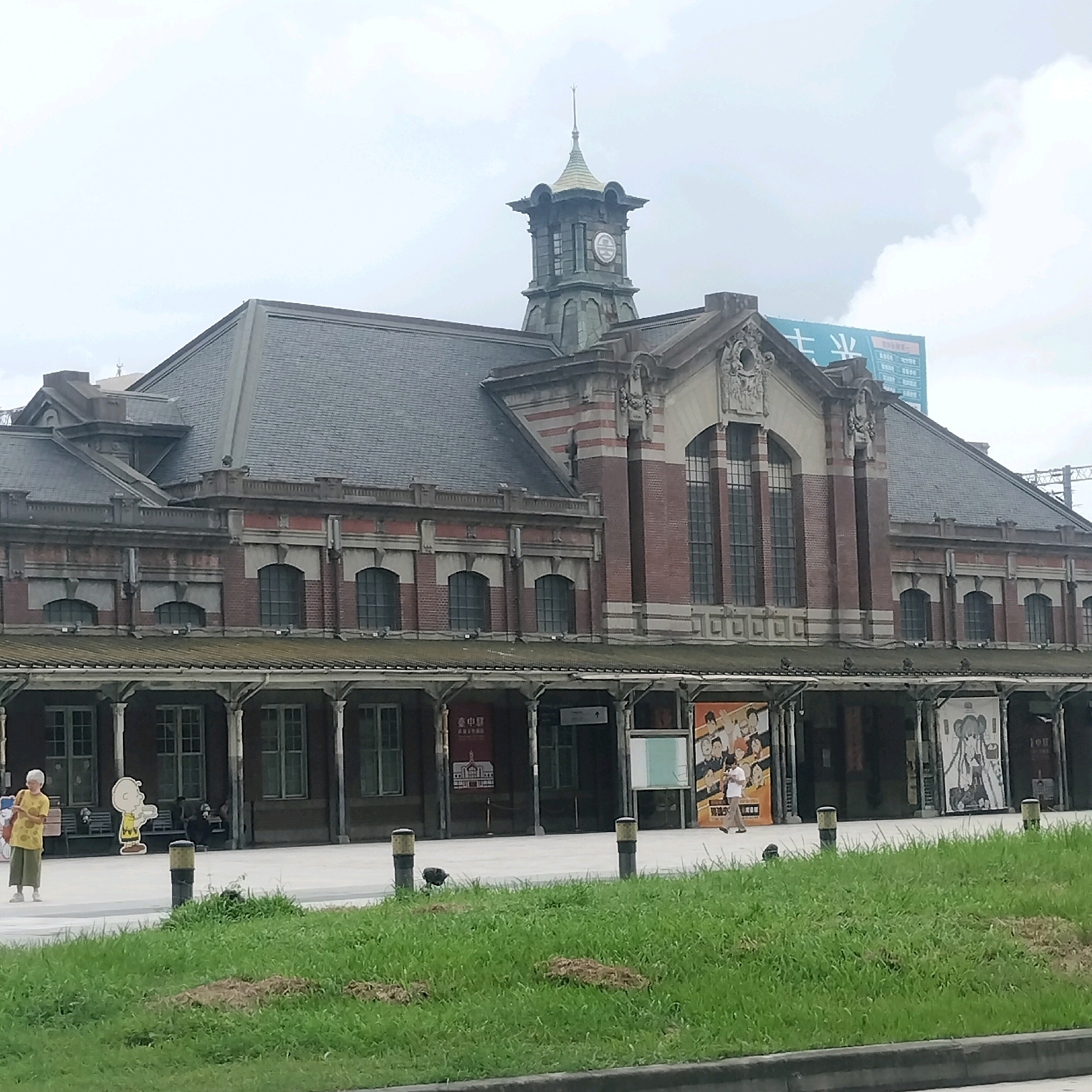The image depicts the exterior of the Taichung Station in Taichung, Taiwan. The building is a grand, historic structure with red brick and stone accents, featuring a prominent clock tower. In front of the station, a grassy area borders a sidewalk where several people are visible. To the left, a woman in a yellow shirt and green pants stands with a bag. Further along the sidewalk, a cutout of the character Charlie Brown is placed. Various posters and banners are displayed along the station's facade, including one featuring anime-style characters. The sky is overcast.
