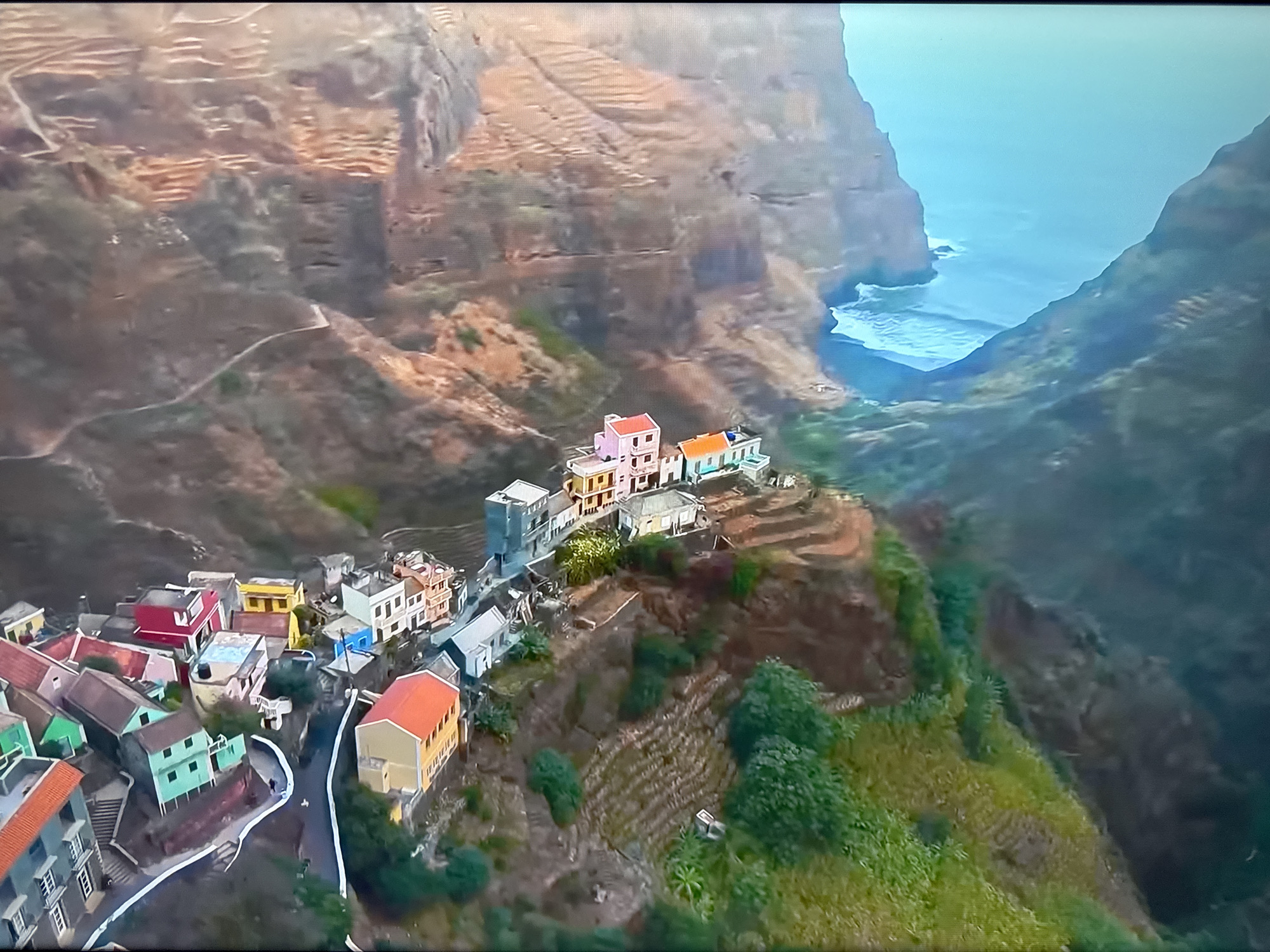 This is an aerial view of a village nestled on steep, terraced hillsides overlooking the ocean. The village consists of numerous brightly colored houses with red, yellow, pink, green, and blue facades, clustered together along winding paths. The surrounding landscape is characterized by rugged, rocky cliffs and lush green vegetation, with cultivated terraces visible on the mountainside. The ocean appears a vibrant blue, with waves crashing against the base of the cliffs. The image depicts a remote, scenic settlement in a dramatic natural environment. There is no indication of the city of Ləcət, Azerbaijan, in this image.