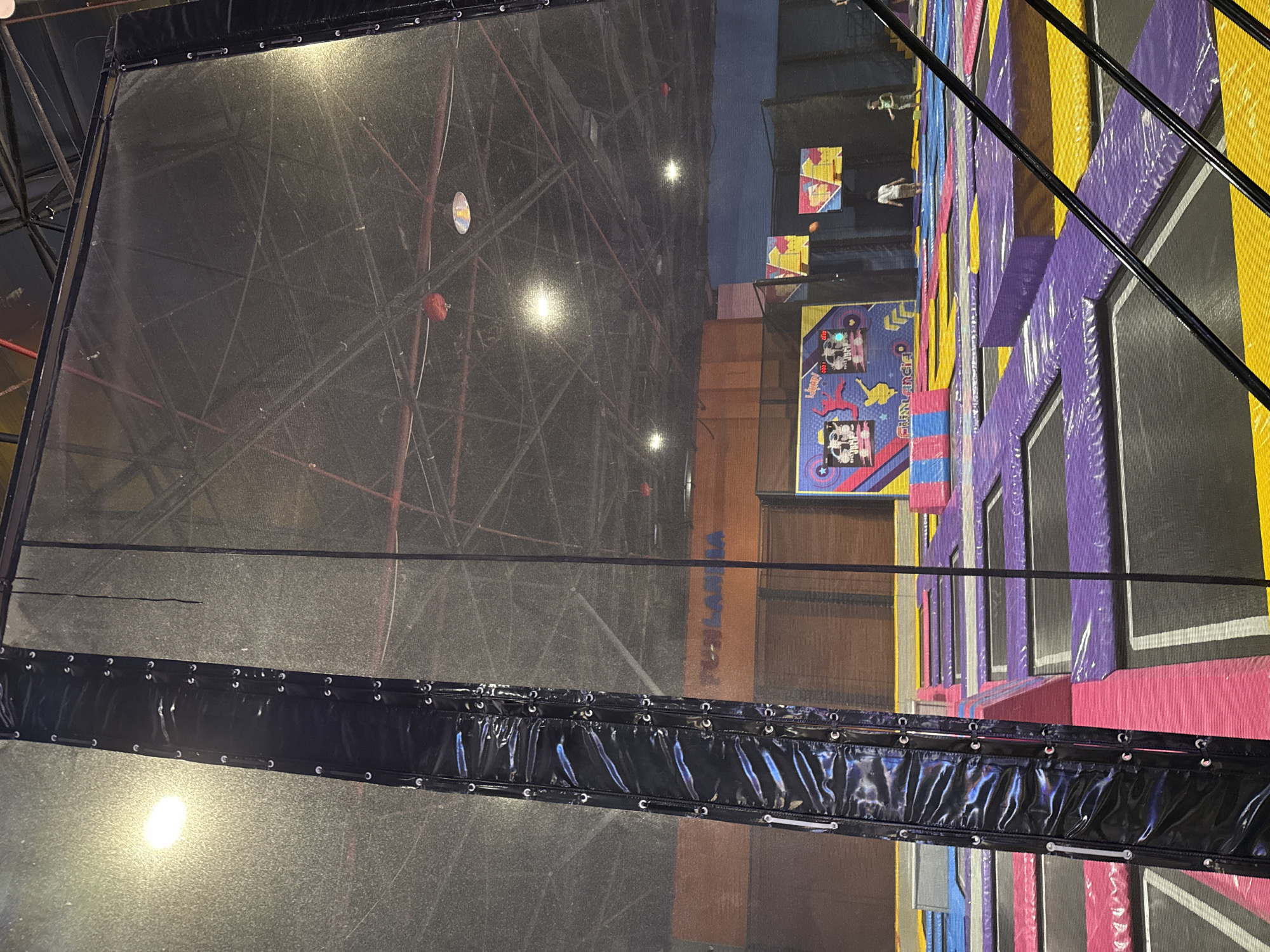The image shows the interior of what appears to be an entertainment venue in Tashkent, Uzbekistan, possibly a trampoline park. In the foreground is a large net enclosure with a black, glossy border, suggesting safety barriers around the activity area. The background features a trampoline area with various colored padded sections, including purple, yellow, and pink, and play structures. Some blurred figures are visible on the trampolines, indicating people engaged in recreational activities. The overall environment is indoors with artificial lighting.