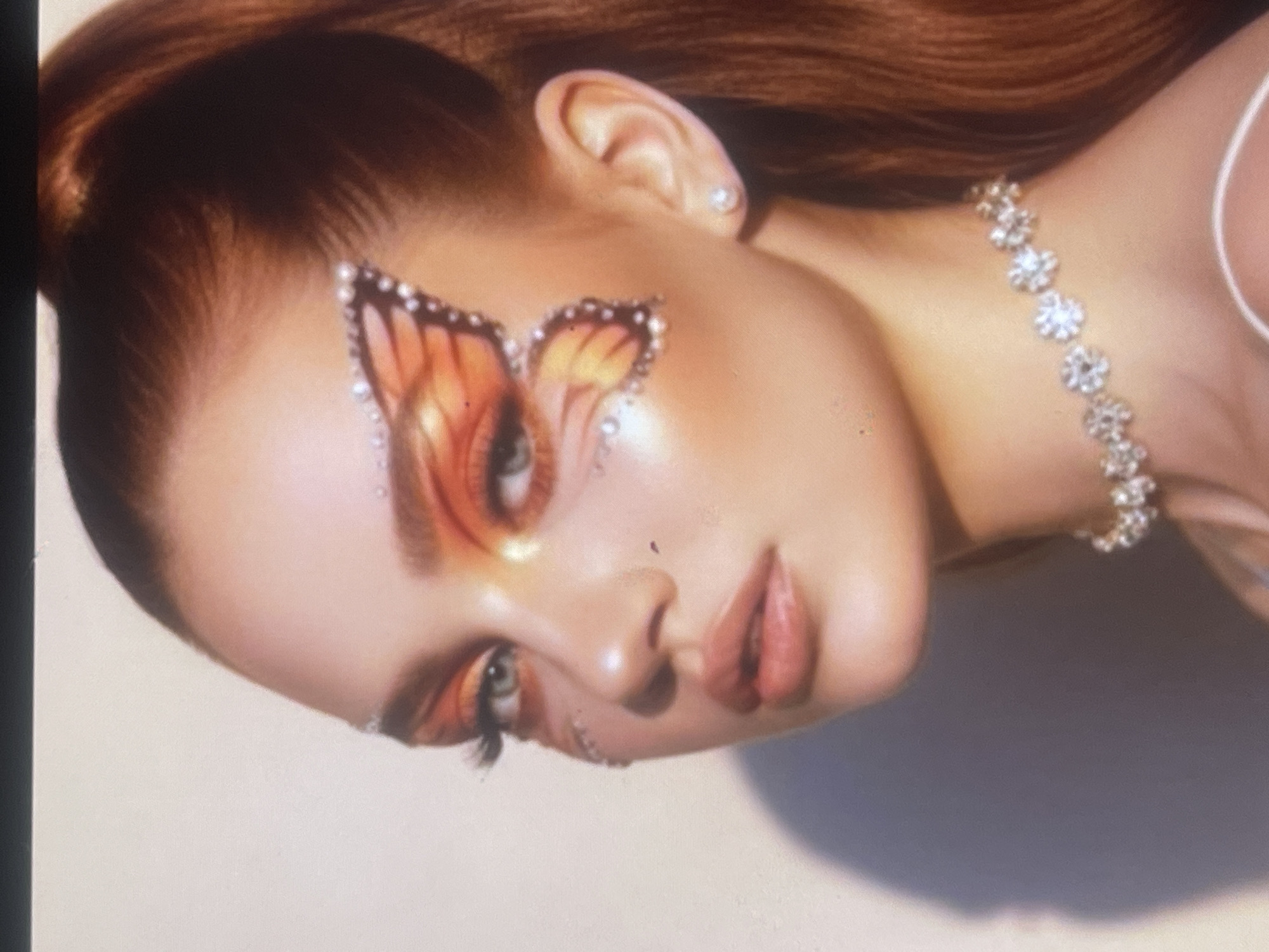 The media file features a close-up portrait of a female individual, her head and upper chest visible, set against a two-tone background that shifts from lighter on the left to darker on the right. The image is tilted counter-clockwise.
The subject is a young woman with reddish-brown hair, styled back from her face. Her skin appears smooth and luminous. The focal point of the image is her elaborate makeup, particularly around her right eye (viewer's right). A vibrant butterfly-wing design, predominantly in shades of orange and yellow with darker accents, is painted around and extending from her eye onto her cheek. This butterfly makeup is intricately outlined with small, sparkling rhinestones or glitter, which also extend below her lower lash line and onto the bridge of her nose. Her lips are a soft, pinkish-nude color.
She wears a sparkling choker-style necklace composed of interconnected, flower-like metallic or jeweled elements on her left side of the neck (viewer's left), and a small, clear stud earring in her visible ear. The woman has a neutral to serene expression, looking slightly upwards and to the viewer's right with her light-colored eyes.
The setting appears to be a studio with controlled, soft lighting that highlights the intricate details of her makeup and jewelry. There is no visible text in the image. The overall impression is one of artistic expression and refined beauty. This media file is associated with Poltava, Ukraine.