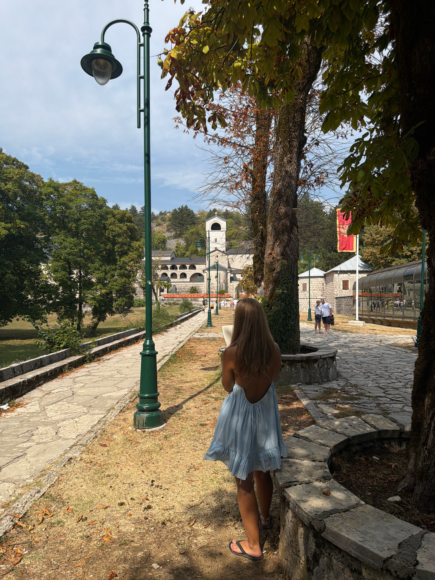 The image depicts the courtyard of the Cetinje Monastery in Cetinje, Montenegro, on a sunny day. A young woman with blonde hair, wearing a light blue dress and flip-flops, stands with her back to the camera, holding a book. She is positioned on a cobblestone path to the right of a tall, dark green lamppost.
The path leads towards a complex of stone buildings with arched windows and a central bell tower, characteristic of the monastery. Several other people are visible in the background, walking and interacting. A large, red banner with a gold emblem hangs from a flagpole on the right side of the courtyard.
The courtyard is lined with stone walls and features several mature trees, their leaves casting shadows on the ground. The ground cover varies between dry grass and fallen leaves. The overall atmosphere suggests a peaceful, historical setting.