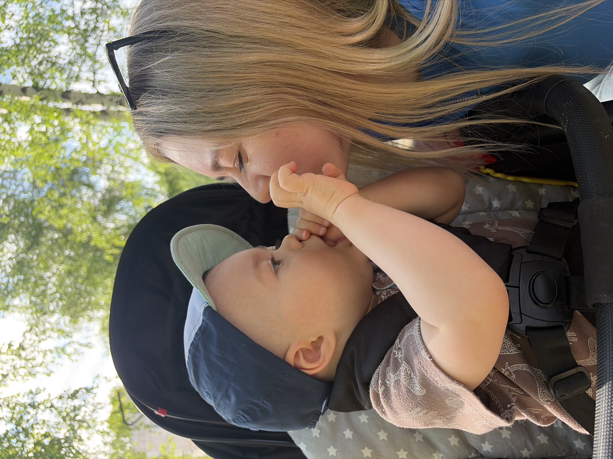 A woman with long blonde hair, wearing black glasses, and a blue shirt is kissing a baby in a stroller. The baby is wearing a navy blue and light green baseball cap, a black shirt, and a brown patterned outfit. The stroller has a black frame, a grey liner with white stars, and a black harness. The background shows trees with green leaves, suggesting an outdoor location, possibly in a park or garden. The image was taken from a low angle, looking up at the woman and baby. There is no definitive evidence to place the image in the city of Chaikovsky, Russia.