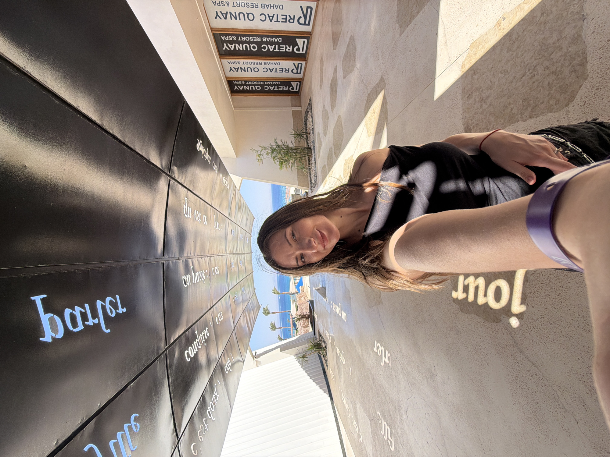 A woman is taking a selfie in an outdoor walkway in Dahab, Egypt. The walkway has a black, curved ceiling with white and blue lettering. The ground is paved with light-colored tiles and also has lettering on it. In the background, palm trees, the ocean, and buildings are visible. Signs for "RETAC QUNAY DAHAB RESORT & SPA" are mounted on a building to the right of the walkway. The woman is wearing a black off-the-shoulder top and has a red string bracelet on her wrist.