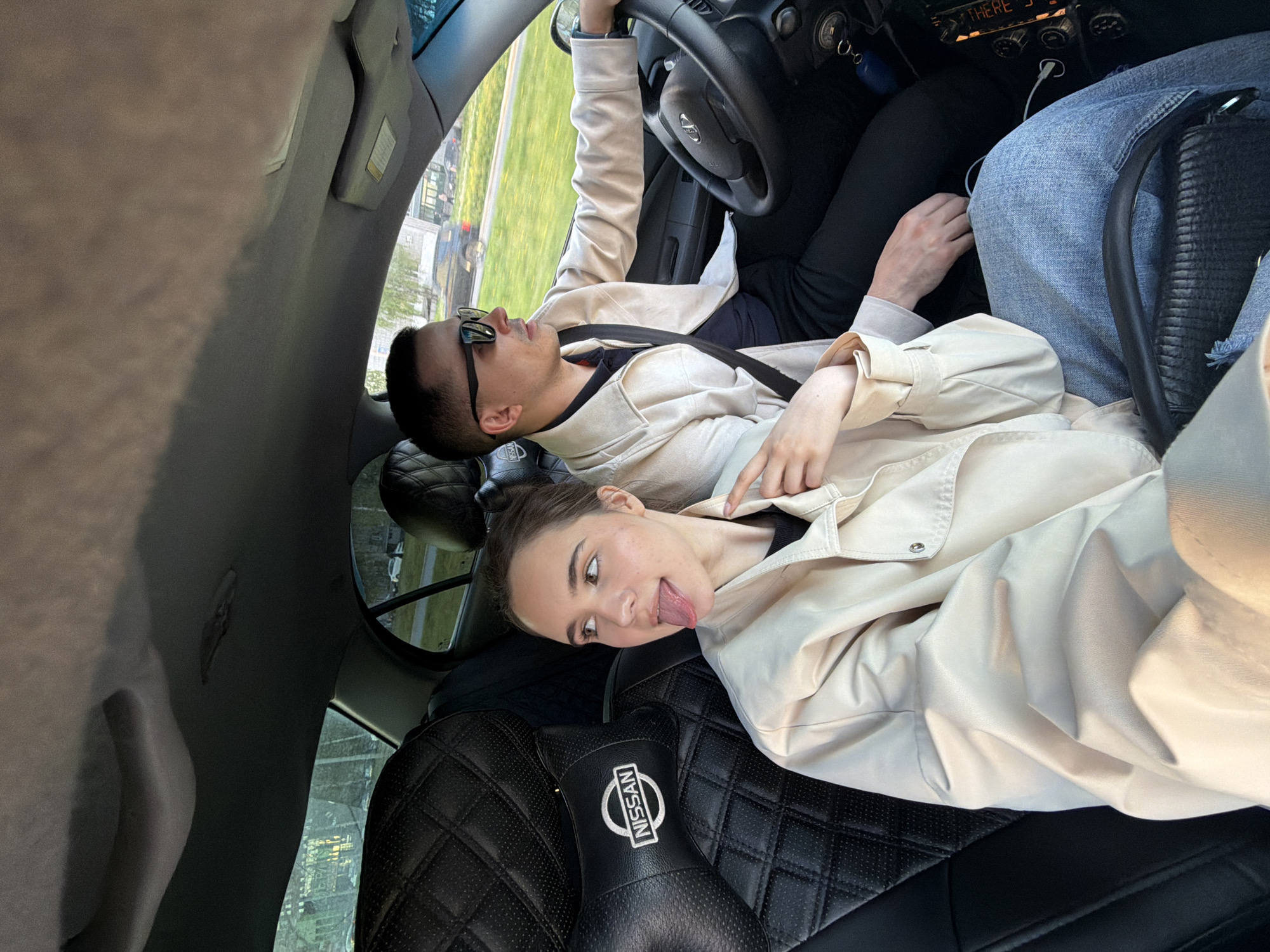 Inside a Nissan vehicle, two individuals are seen. The man, wearing sunglasses and a beige jacket, is at the wheel. The woman, also in a beige jacket, is in the passenger seat, sticking her tongue out. The car's interior features black leather seats with a quilted pattern and a Nissan logo on the headrest. Through the windows, a blurred urban environment is visible. The location cannot be determined to be Izhevsk, Russia.