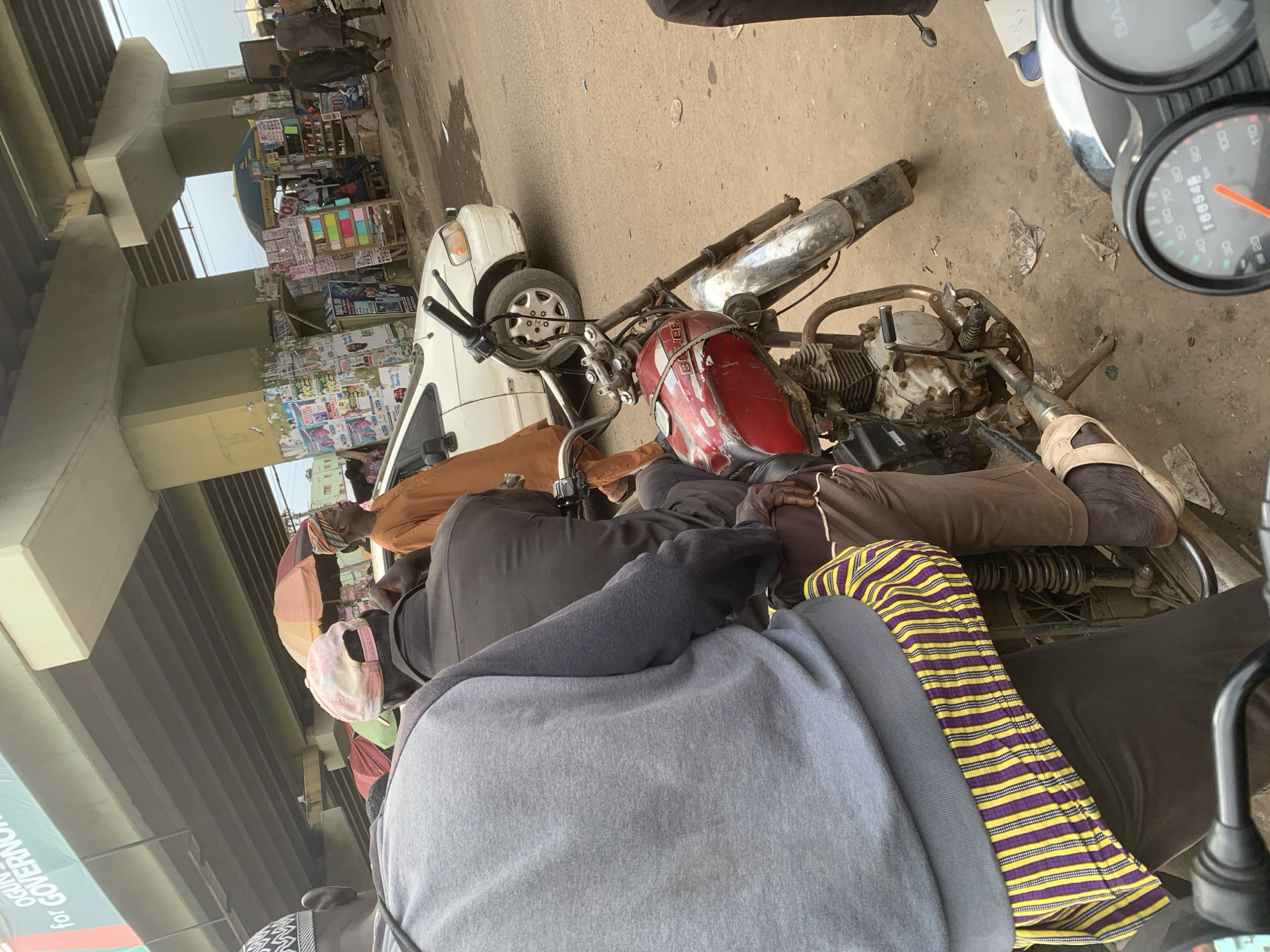 In Shagamu, Nigeria, beneath an elevated highway, a group of people are gathered on a street. The foreground is dominated by the rear view of a motorcycle with a red fuel tank and various chrome components. A person wearing a gray sweatshirt and a colorful striped garment is seated on the motorcycle. Behind them, another individual in a dark shirt is also on the motorcycle. A white car is visible in the background, along with market stalls and a few other pedestrians. The environment suggests a busy urban setting with signs of commercial activity and traffic.