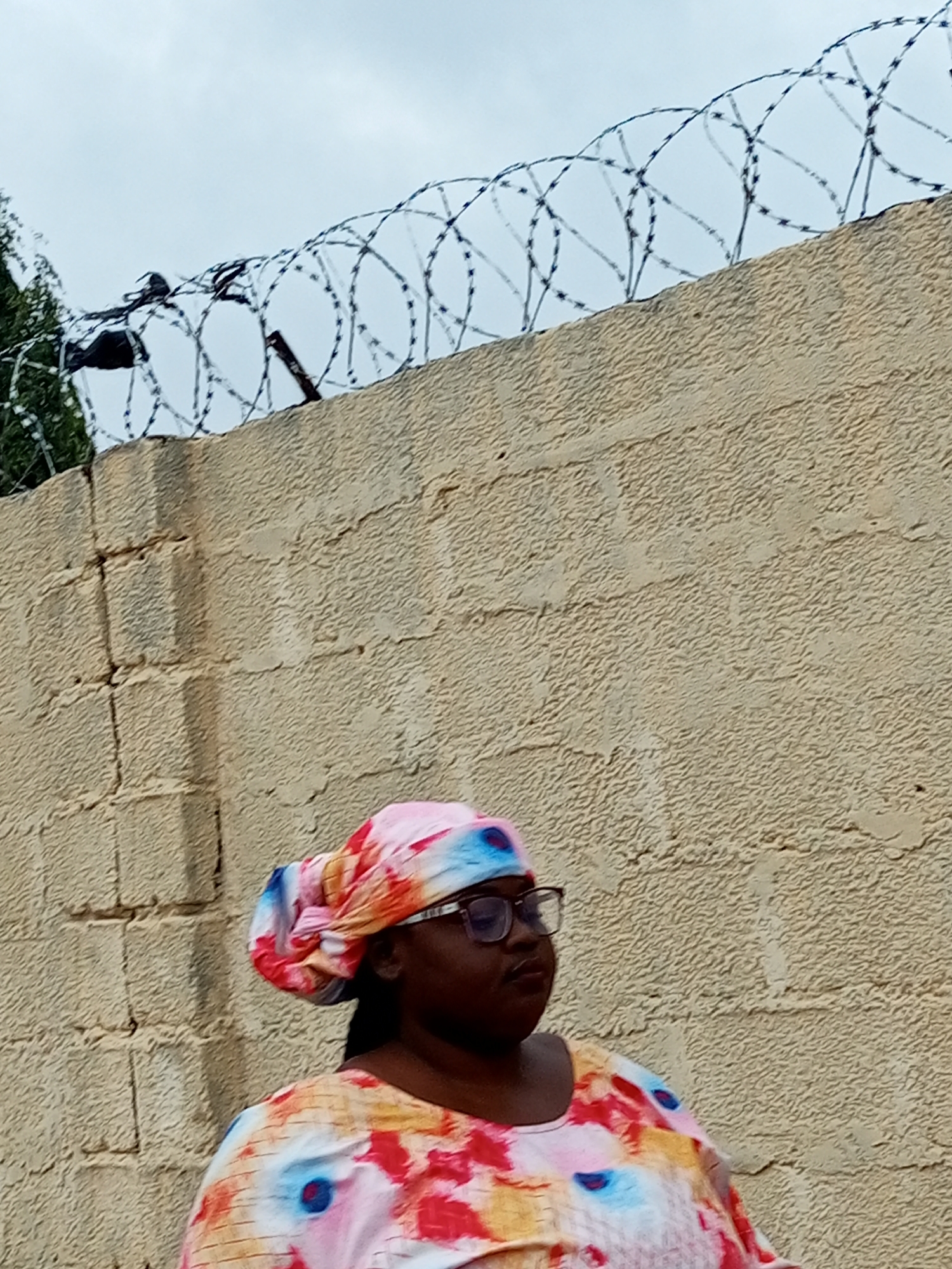 In an outdoor, urban setting under an overcast sky, a person wearing glasses, a colorful headscarf, and a matching brightly patterned garment is positioned against a light brown textured wall. Barbed wire is visible along the top of the wall, and to the left of the frame, a dense green foliage is present. The wall appears to be constructed of large, rectangular blocks. The person is facing towards the right of the image with their head slightly tilted down.