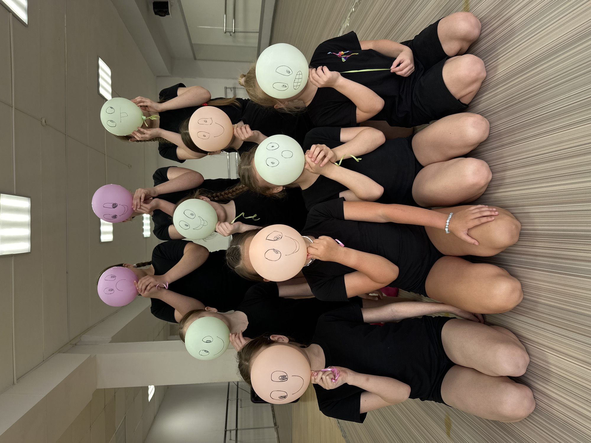 A group of approximately ten young individuals, appearing to be children or pre-teens, are arranged in diagonal rows on a light-colored, striped floor within an indoor setting. They are uniformly dressed in black t-shirts and shorts. Each person holds a pastel-colored balloon in front of their face, effectively obscuring their features. The balloons are in shades of light green, pink, and light orange/beige, and each features a unique, hand-drawn face (smiley, silly, or surprised) created with a black marker. Small ribbons are visible on some of the balloons.
The individuals are sitting or kneeling closely together, engaged in a playful group activity, possibly a photo session, a class exercise, or a performance. The setting is a bright, spacious room with plain light-colored walls and overhead fluorescent lighting panels, characteristic of a studio, gymnasium, or activity hall. The arrangement and balloon faces suggest a lighthearted and joyful atmosphere. No discernible text is visible within the image. This scene takes place in Votkinsk, Russia.