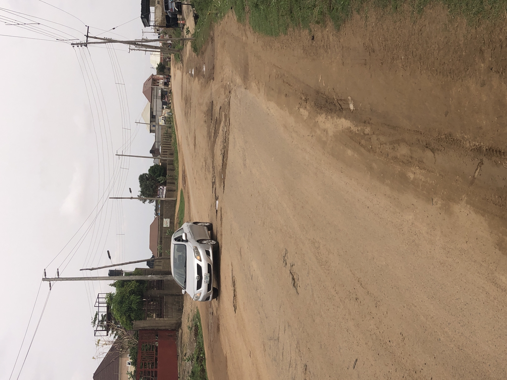 A silver car is parked on a dirt road in what appears to be a residential area in Hanwa, Nigeria. The road is unpaved and contains potholes. There are utility poles with power lines along the roadside. Houses and vegetation are visible in the background. The sky is overcast.