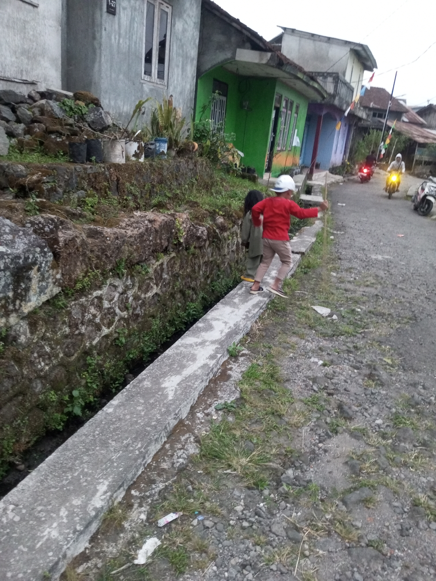 In Andongsili, Indonesia, two children are observed near a concrete drainage channel that runs alongside a dirt road. The child in the foreground, wearing a red shirt and light-colored pants, is facing away from the camera and appears to be in motion, possibly walking or jumping along the edge of the drainage channel. They are wearing a white helmet. A second child, partially visible behind the first, is wearing a dark top and light-colored bottoms. The environment features a stone wall to the left of the drainage channel, with vegetation growing on it and around its base. The road surface is gravelly. In the background, several houses are visible, painted in various colors including green, blue, and purple, with some bearing decorative flags. Two motorcycles are present on the road, one ridden by a person in a white garment. The sky is overcast, suggesting a diffused light.