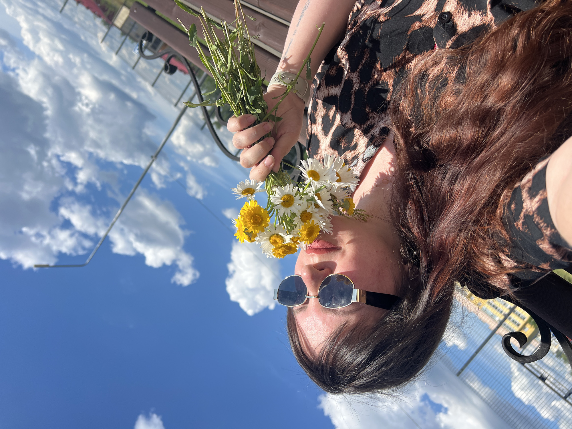 A young woman with long, dark brown hair and a leopard-print top is sitting on a bench outdoors, holding a bouquet of white and yellow daisies up to her face, possibly smelling them. She is wearing sunglasses with silver and black frames. The background shows a bright blue sky with white, fluffy clouds, and a faint outline of buildings and a fence. A street lamp is visible in the upper right portion of the image. The scene appears to be set in Noviy, Russia, suggesting a public park or an outdoor recreational area given the presence of benches and a fence. The lighting indicates it is daytime, likely late afternoon or early evening due to the angle of the sun casting some shadows.
