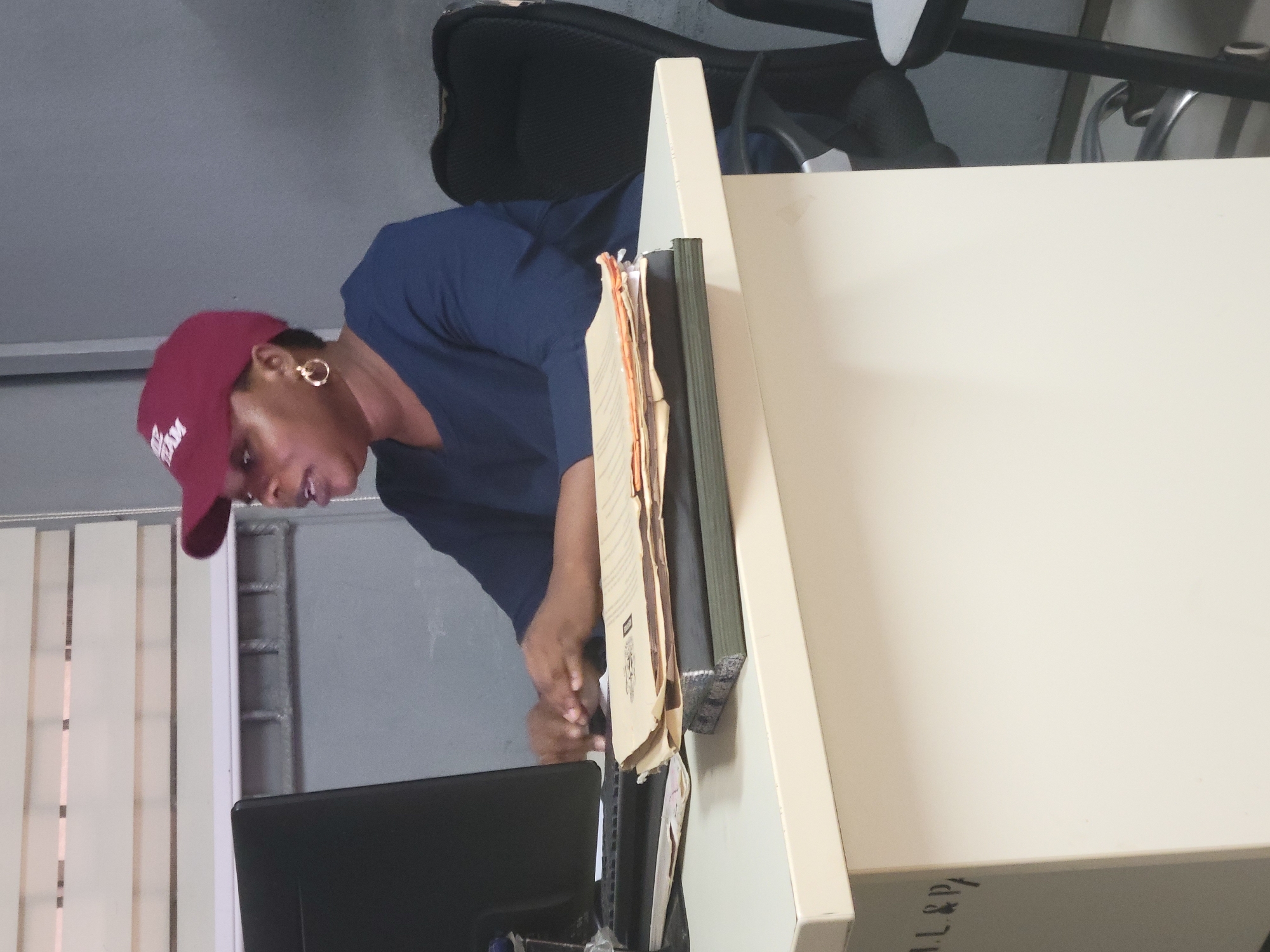 An indoor, vertical-format image captures a person leaning over a desk, engaged in work within what appears to be an office setting in Abuja, Nigeria.
The main subject is an individual, likely female, with dark skin, wearing a dark blue short-sleeved top and a maroon or burgundy baseball cap with white text that appears to read "ABAM". She wears a gold-colored hoop earring on her visible ear. Her head is tilted downwards, and her expression suggests focus or attention to tasks on the desk.
The setting is a professional workspace, likely an office cubicle or desk area. A light-colored (beige or off-white) desk or partition dominates the right side of the frame. On the desk, several paper files and folders are stacked, some appearing well-used. A black computer monitor is visible in the lower left, towards which the person is looking. A black ergonomic office chair is partially visible in the background behind the desk. To the far left, vertical window blinds are present, and a grey wall or partition with some metal fixtures is visible behind the person.
The activity taking place is desk work, possibly reviewing documents or interacting with the computer. The person's hands are near the files and monitor, indicating active engagement.
Notable details include the casual but focused attire, the array of physical files suggesting administrative or record-keeping work, and the general ambience of a busy office environment. Visible text includes "ABAM" on the baseball cap and partially visible handwritten text "AL&P" on the edge of the desk. The time of day cannot be determined, but the lighting suggests typical indoor conditions.