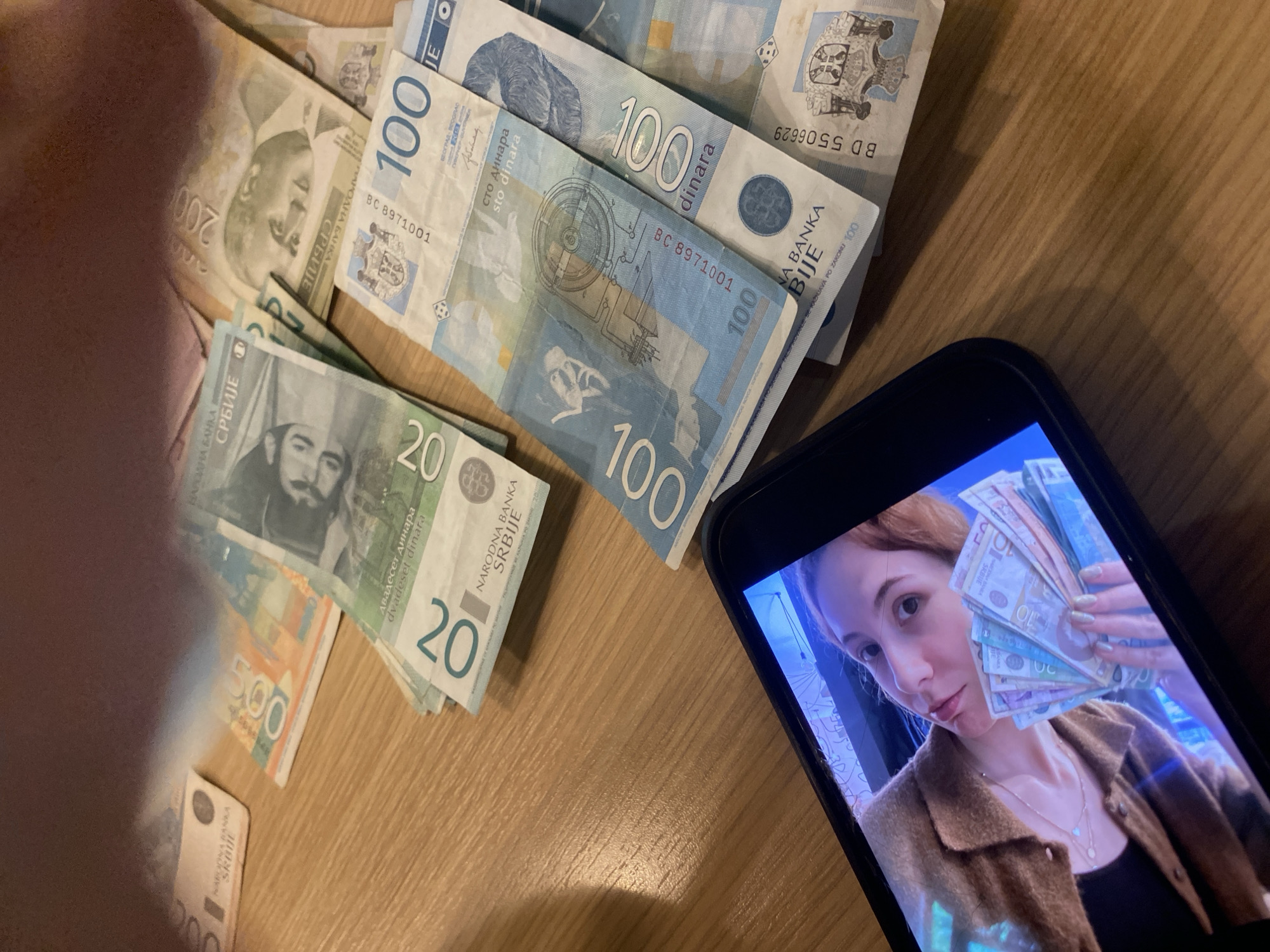 A tabletop contains several Serbian Dinar banknotes (100, 20, 200, and 500) along with a cell phone displaying a video of a woman holding Serbian Dinar banknotes. The table is made of wood. The location in the city of Novi Sad, Serbia is not identifiable.