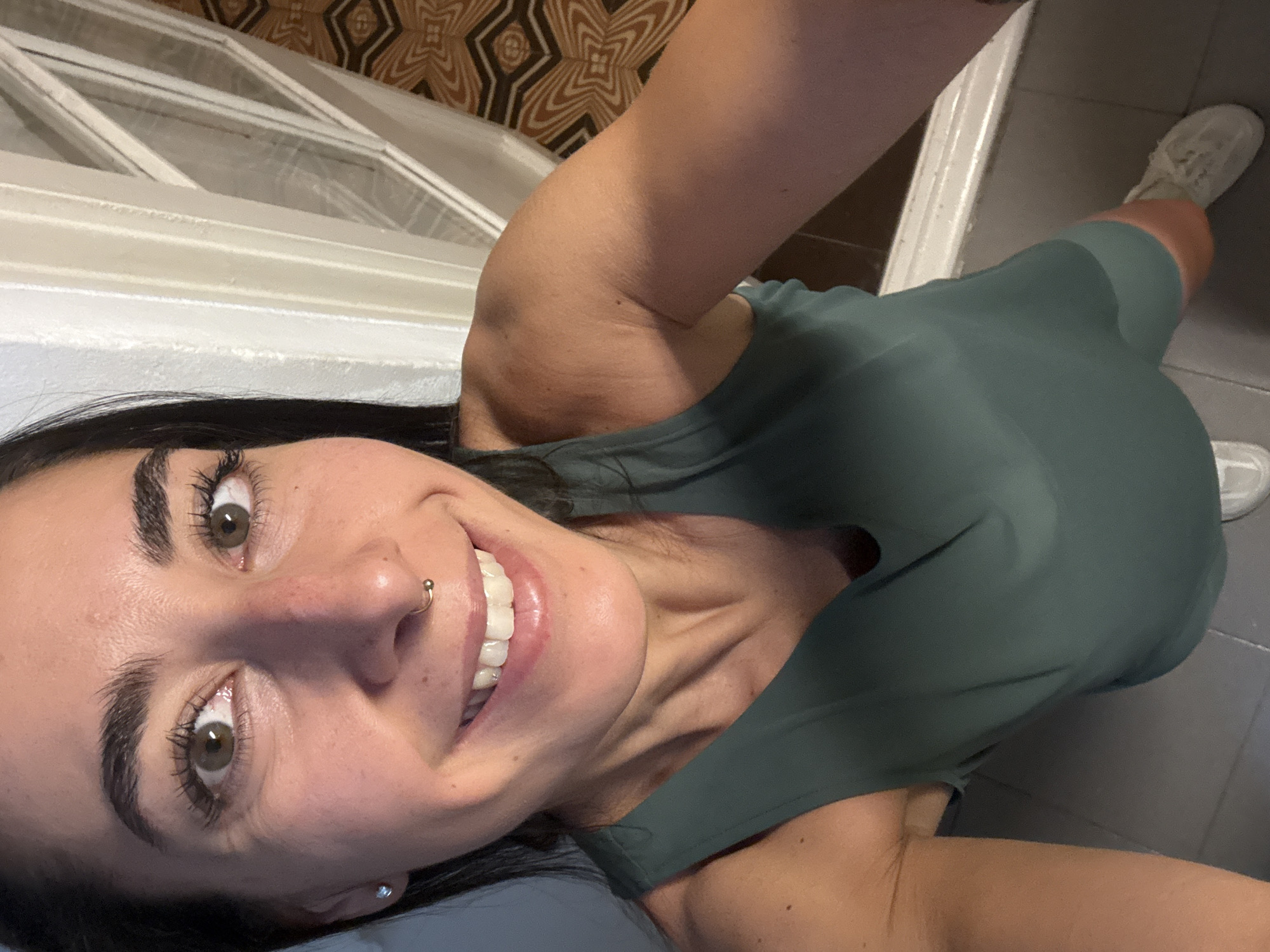 The image is a high-angle, first-person perspective selfie of a woman with dark hair and brown eyes. She is wearing a dark green tank top and white sneakers. She has a nose ring and a small earring in her left ear. She is smiling broadly, revealing her teeth. The background includes a white door frame or window frame, a patterned curtain or wallpaper, and a tiled floor. The scene appears to be indoors, possibly in a home or hotel room, and is not identifiable as being in the city of Barcelona, Spain.