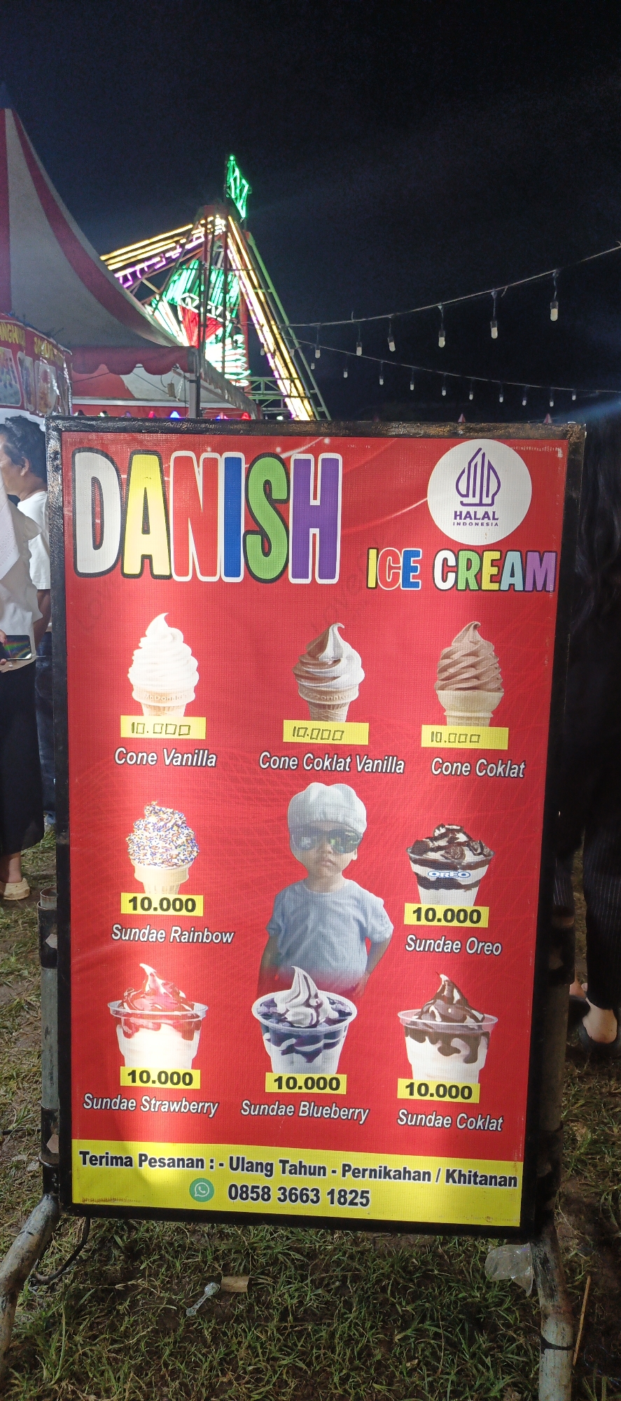 This is a photograph of an outdoor ice cream stand at night, likely in Banding Agung, Indonesia, given the Indonesian text on the sign. The focal point is a large, red banner advertising "DANISH ICE CREAM". The banner displays various ice cream flavors and sundaes with prices listed in Indonesian Rupiah. Prominently featured are images of cones with vanilla and chocolate ice cream, and sundaes with rainbow sprinkles, strawberry, blueberry, and Oreo toppings. The price for most items appears to be 10,000 Rupiah. In the center of the banner, a young child wearing a cap and sunglasses is depicted. At the bottom of the banner, contact information for placing orders is provided, including a phone number and a WhatsApp icon. In the background, elements of a fairground or festival are visible, including colorful lights and tent structures. The ground is covered with grass. The overall scene suggests a lively, festive atmosphere associated with a local event or market.