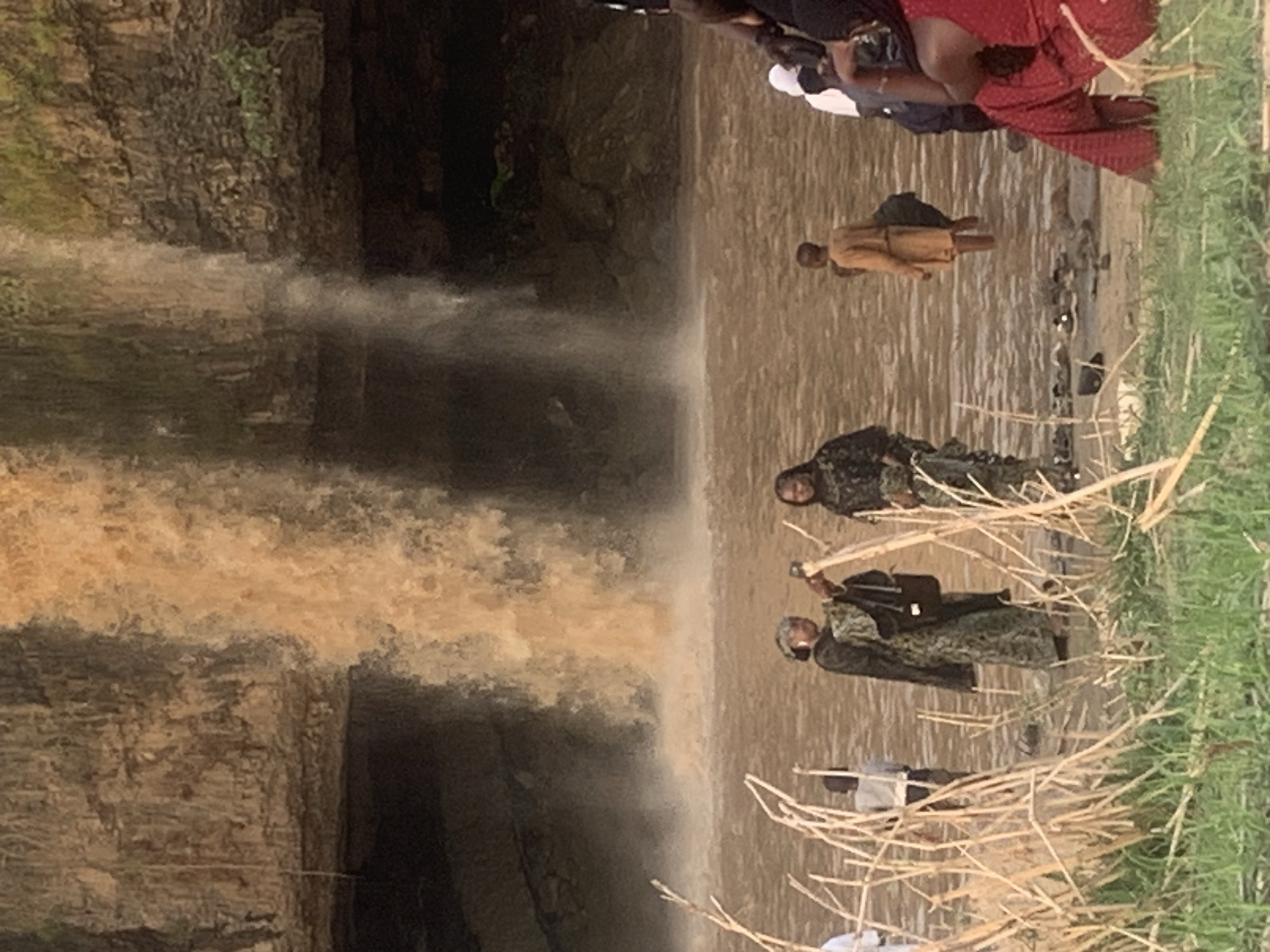 The image shows Gurara Waterfalls in Kukum, Nigeria. The waterfall has muddy water flowing from a rocky cliff into a shallow pool. Several people are visible: one standing alone in the pool, a group standing in the water at the edge of the pool, and two individuals standing on the grass at the edge of the pool, near dried reeds.