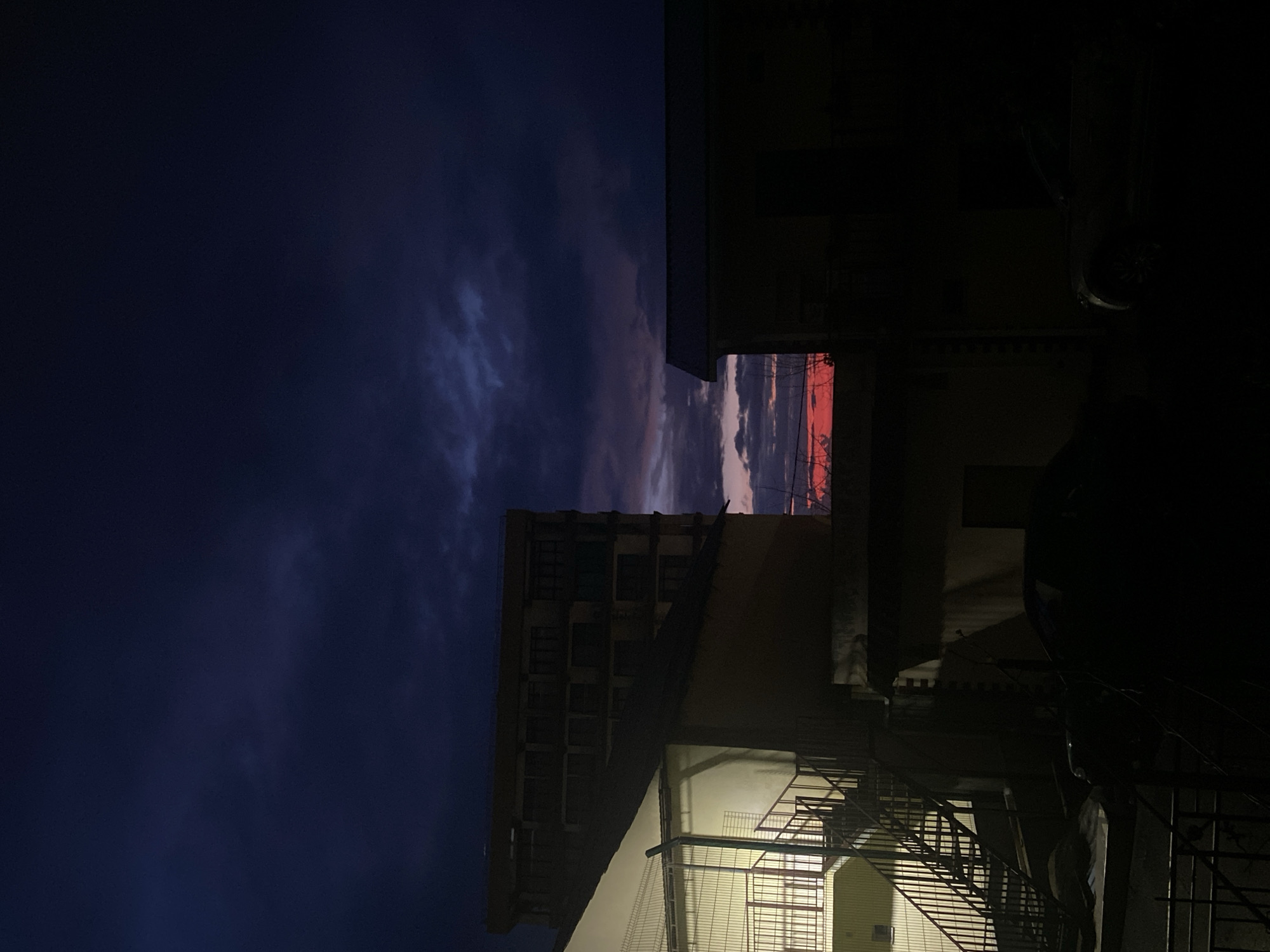 The image is an upward view of buildings at dusk or night. A dark blue sky fills the left side of the frame, with faint clouds visible. Buildings occupy the right side of the image, appearing mostly silhouetted due to low light. One building has a fire escape with visible metal stairs. A gap between buildings allows a glimpse of a red and grey sunset-like sky. Due to the lack of identifying features, pinpointing the exact location in Sochi, Russia is not possible. The scene is urban and quiet. No people or specific actions are discernible.