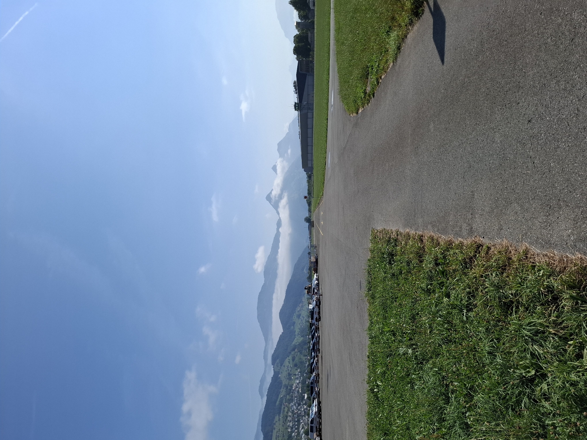 The image depicts a scene at Buochs Airport in Buochs, Switzerland. The foreground shows a paved asphalt surface, likely a taxiway or runway, with a grassy area to the left. Beyond the asphalt, a parking lot is visible, containing numerous cars and vans. In the midground, a large hangar-like building is present, along with a control tower. The background is dominated by a chain of mountains, some of which are partially obscured by clouds, and a clear blue sky above. The overall impression is a serene, daytime view of a small airport surrounded by a mountainous landscape.